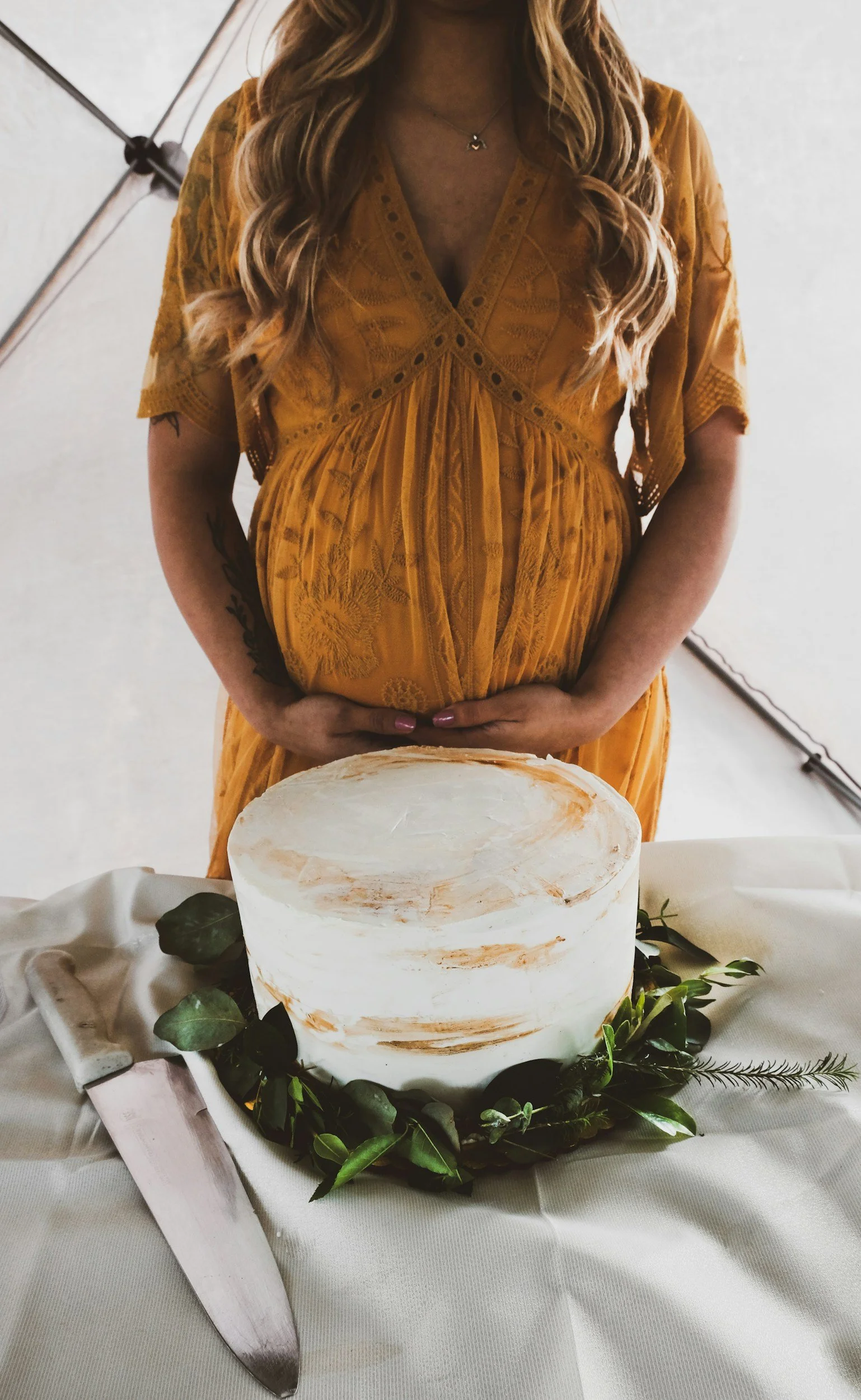 A woman with long wavy hair in a yellow dress standing behind a table with a white cake decorated with green leaves, a large knife, and a beige cloth.