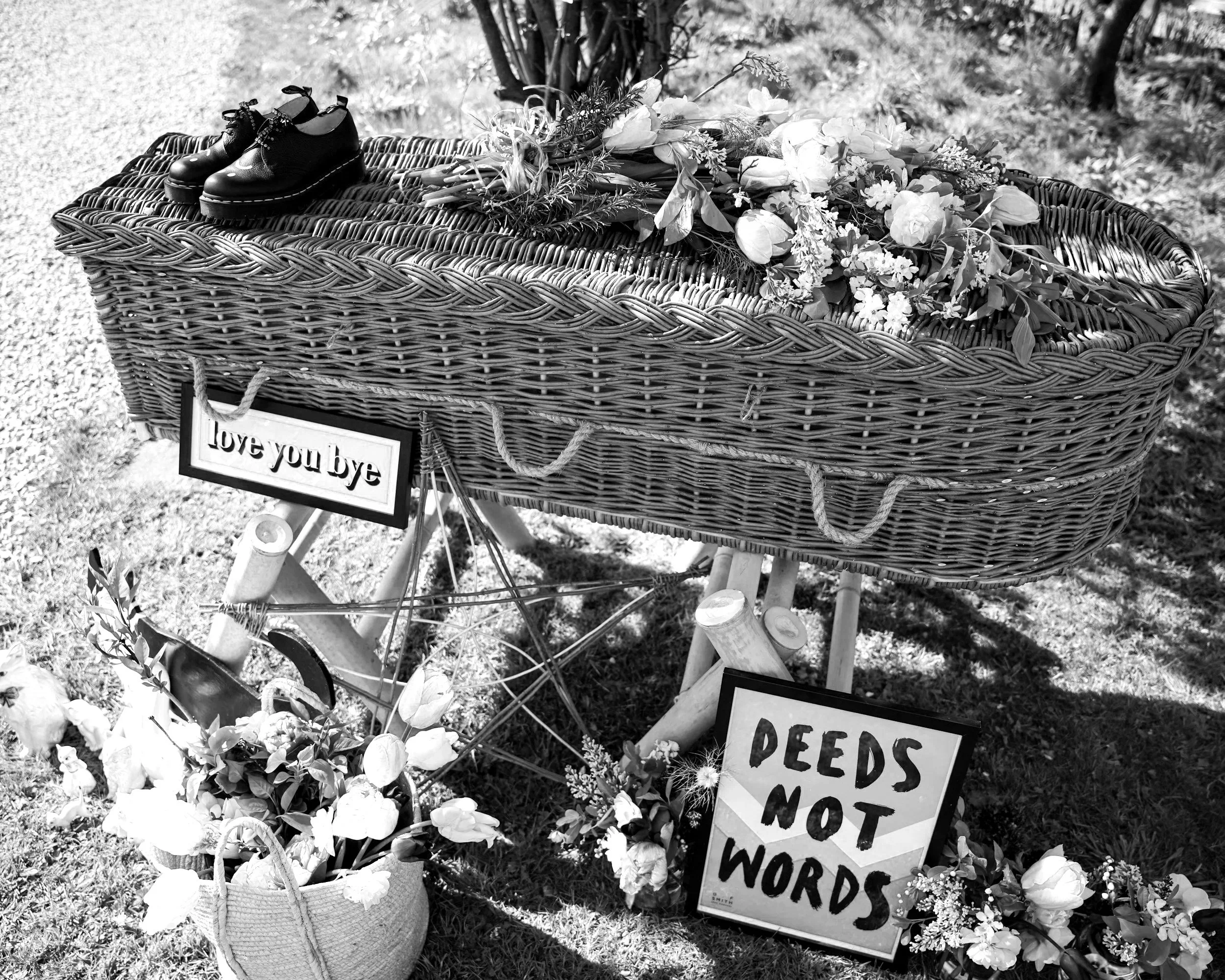 A wicker casket decorated with flowers and greenery, with small shoes on top, placed outdoors. Signs nearby with partially visible text, including "Love you bye" and a sign reading "DEEDS NOT WORDS."