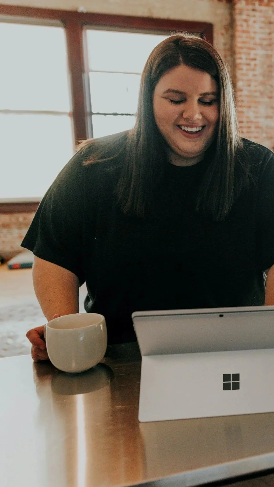 A woman with dark hair and a black shirt sitting at a table, smiling and looking at a tablet, with a white mug in her right hand, against a background of brick walls and large windows.