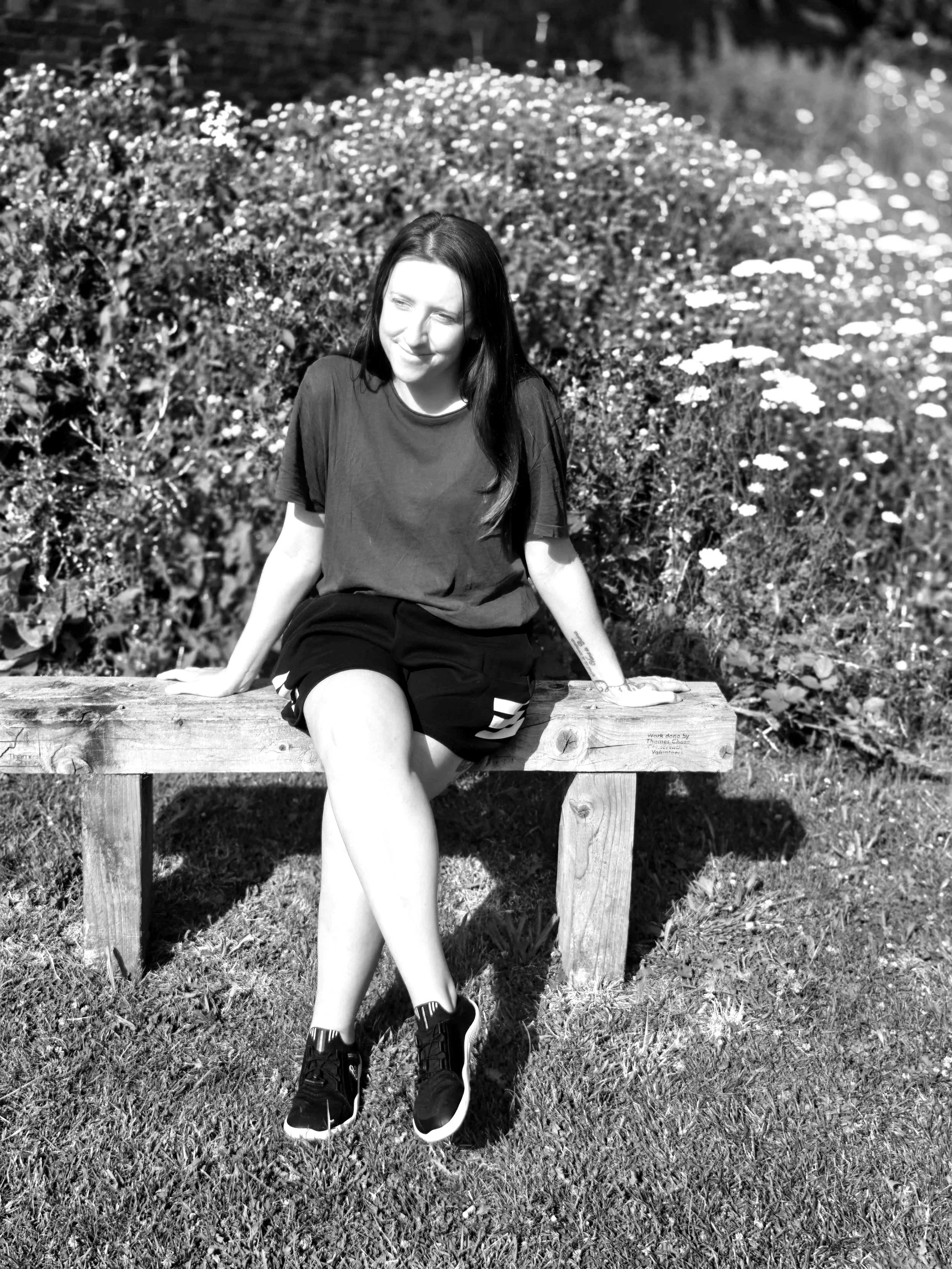 A young woman with long dark hair sitting on a wooden bench outdoors in a garden or park. She is wearing a dark t-shirt, shorts, and sneakers, smiling, with her hands resting on the bench. In the background, there are flowers and foliage, and the photo is in black and white.