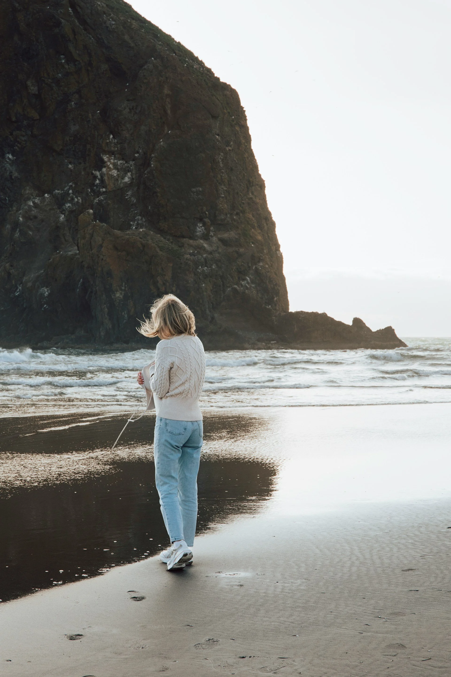 A person with blonde hair walking alone on a beach at sunset, with footprints in the wet sand and a large rock formation in the background.