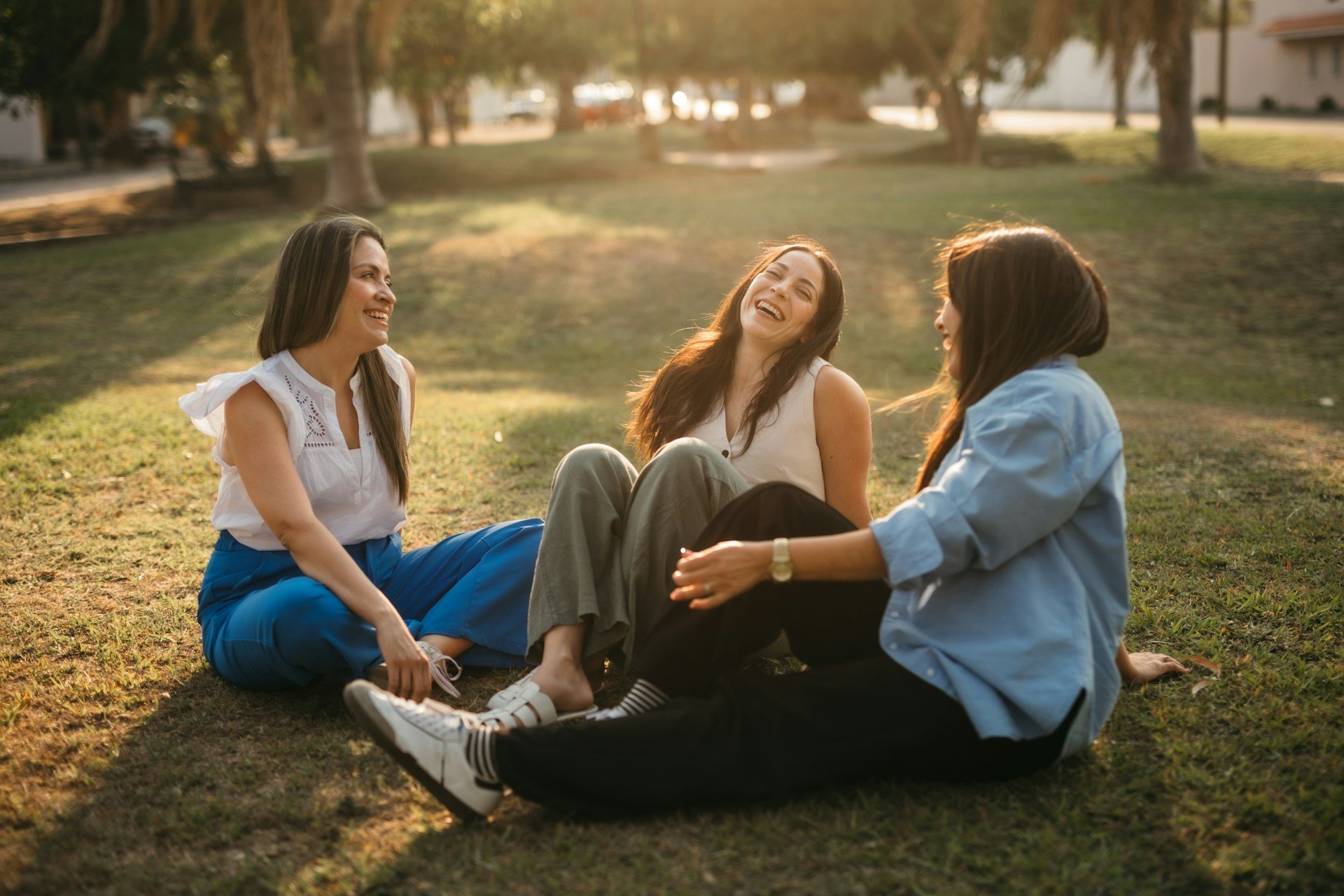 Three women sitting on the grass in a park, smiling and laughing together during sunset.