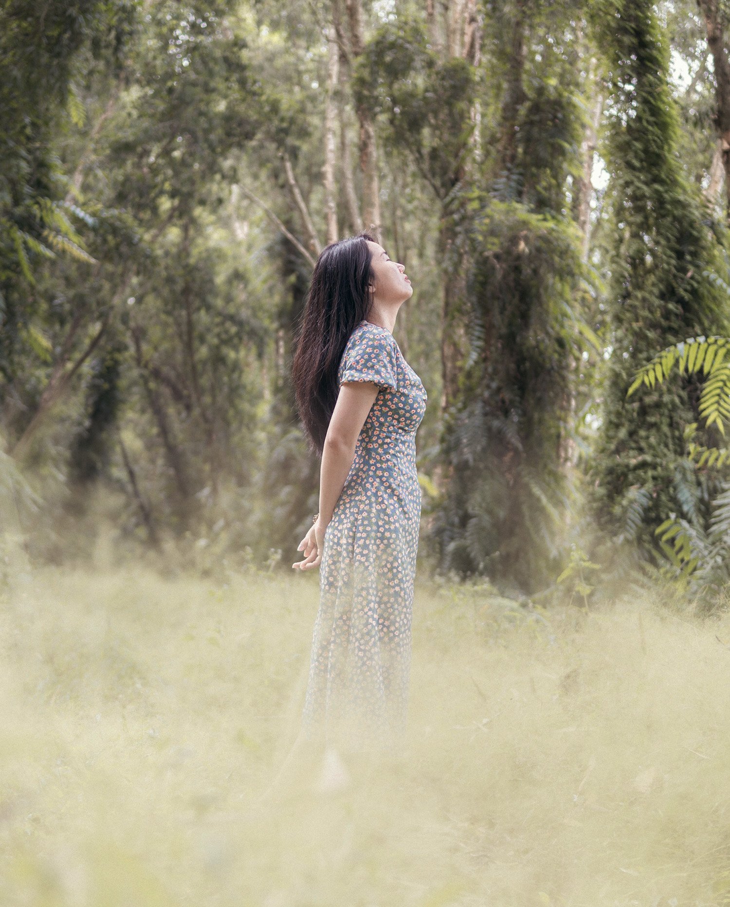 A woman with long dark hair in a floral dress standing outside in a green forest with her face tilted upward and her eyes closed.