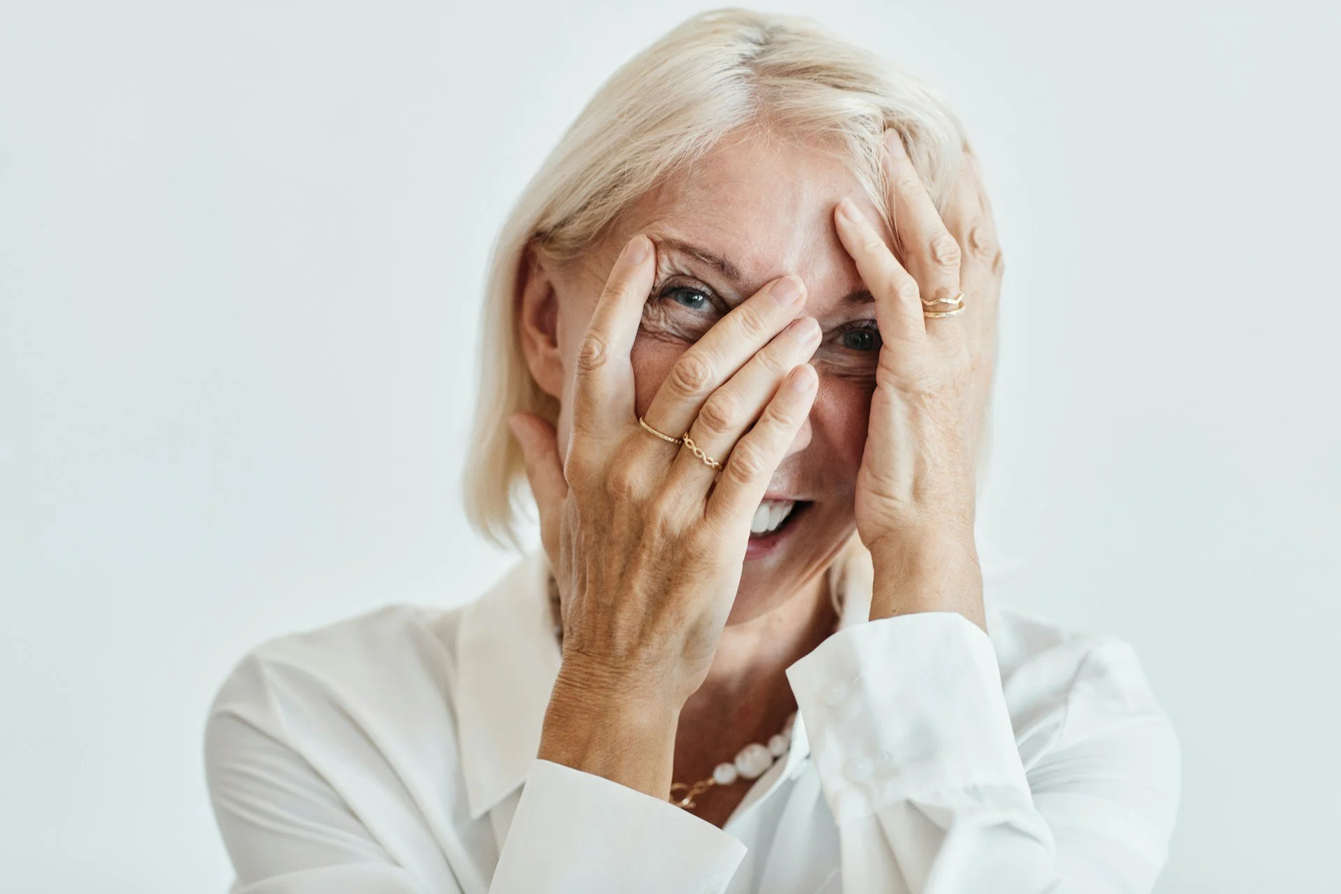 An elderly woman with blonde hair smiling as she covers part of her face with her hands, appearing joyful or amused, against a plain light background.