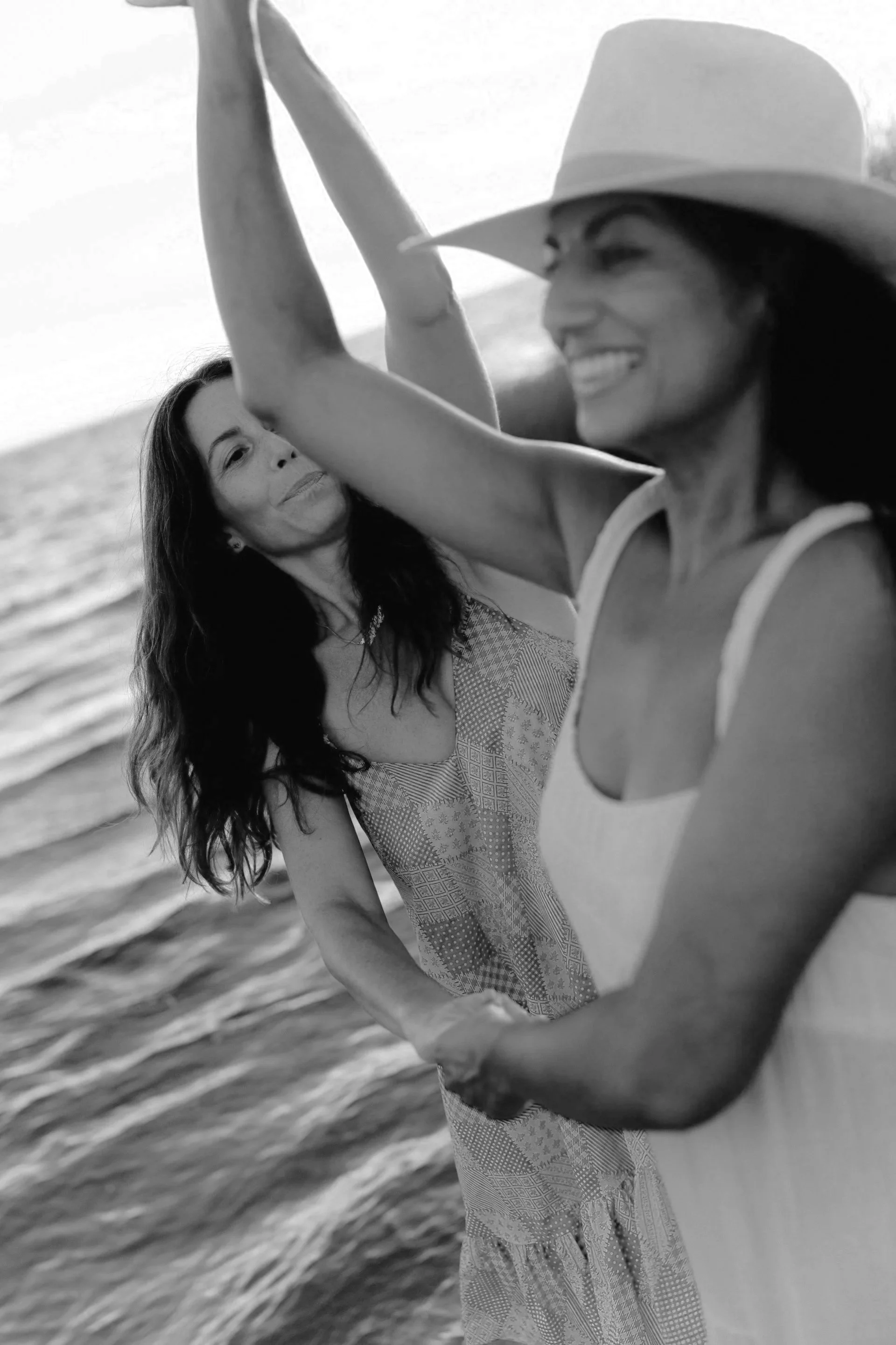 Two women dancing on the beach, one wearing a wide-brimmed hat, smiling and looking at each other, with ocean waves in the background, in black and white.