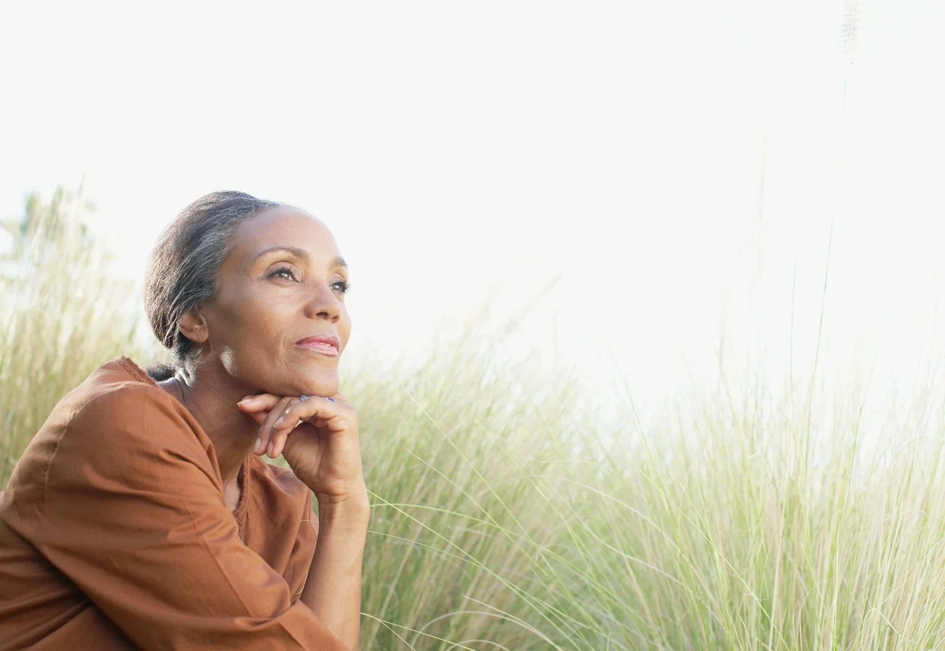 An older woman with short gray hair explores a serene field of tall, green grass during daylight, with a peaceful expression on her face.