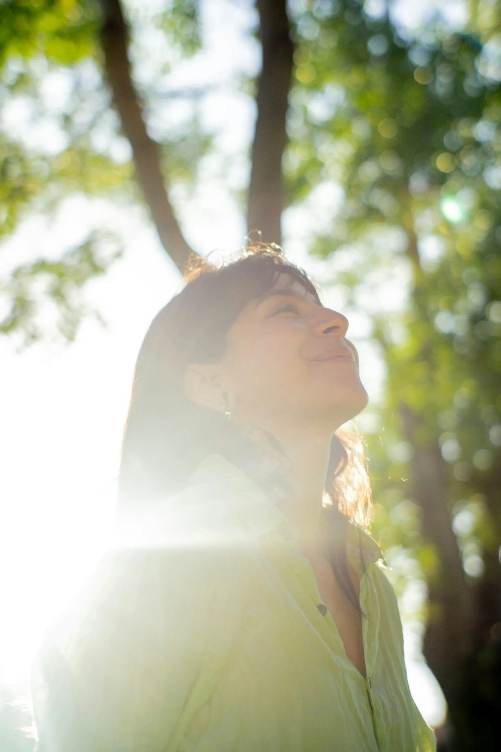 A woman smiling outdoors, with sunlight and trees in the background, her face turned slightly upward.