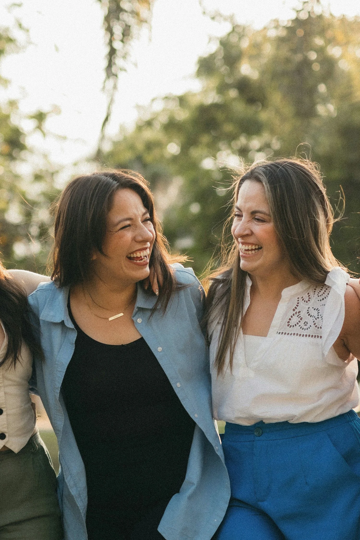 Two women are standing outdoors, smiling and laughing together. They are dressed casually, with one wearing a light blue shirt and the other a white blouse. The background shows trees and sunlight, indicating a warm, sunny day.