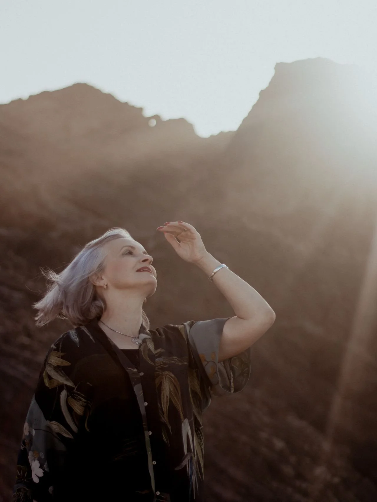 An older woman with gray hair, wearing a patterned blouse, is smiling and raising her hand to shading her eyes while standing outdoors with mountains in the background during sunset or sunrise.