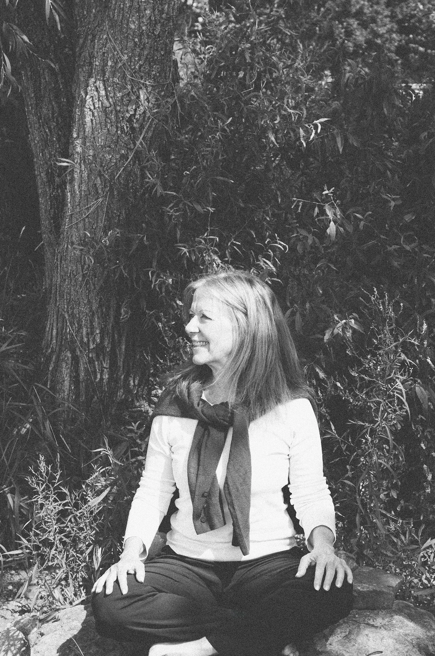 Montreal-based coach Liz Stokvis sitting cross-legged outdoors, smiling, in front of trees and foliage.