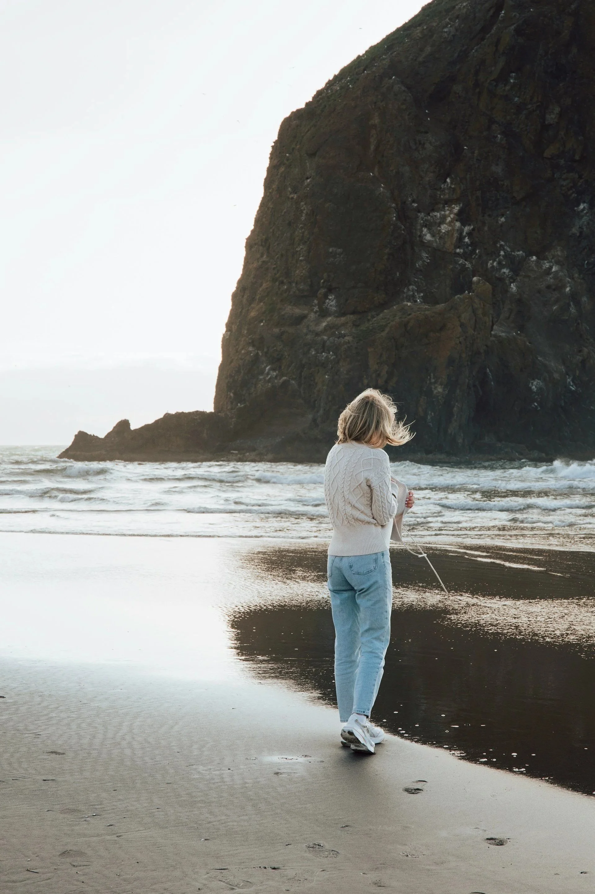A woman with blonde hair, wearing a white sweater and light blue jeans, is walking along the shoreline at the beach with a large rocky cliff in the background. She is holding a string of a kite that is not visible in the image.