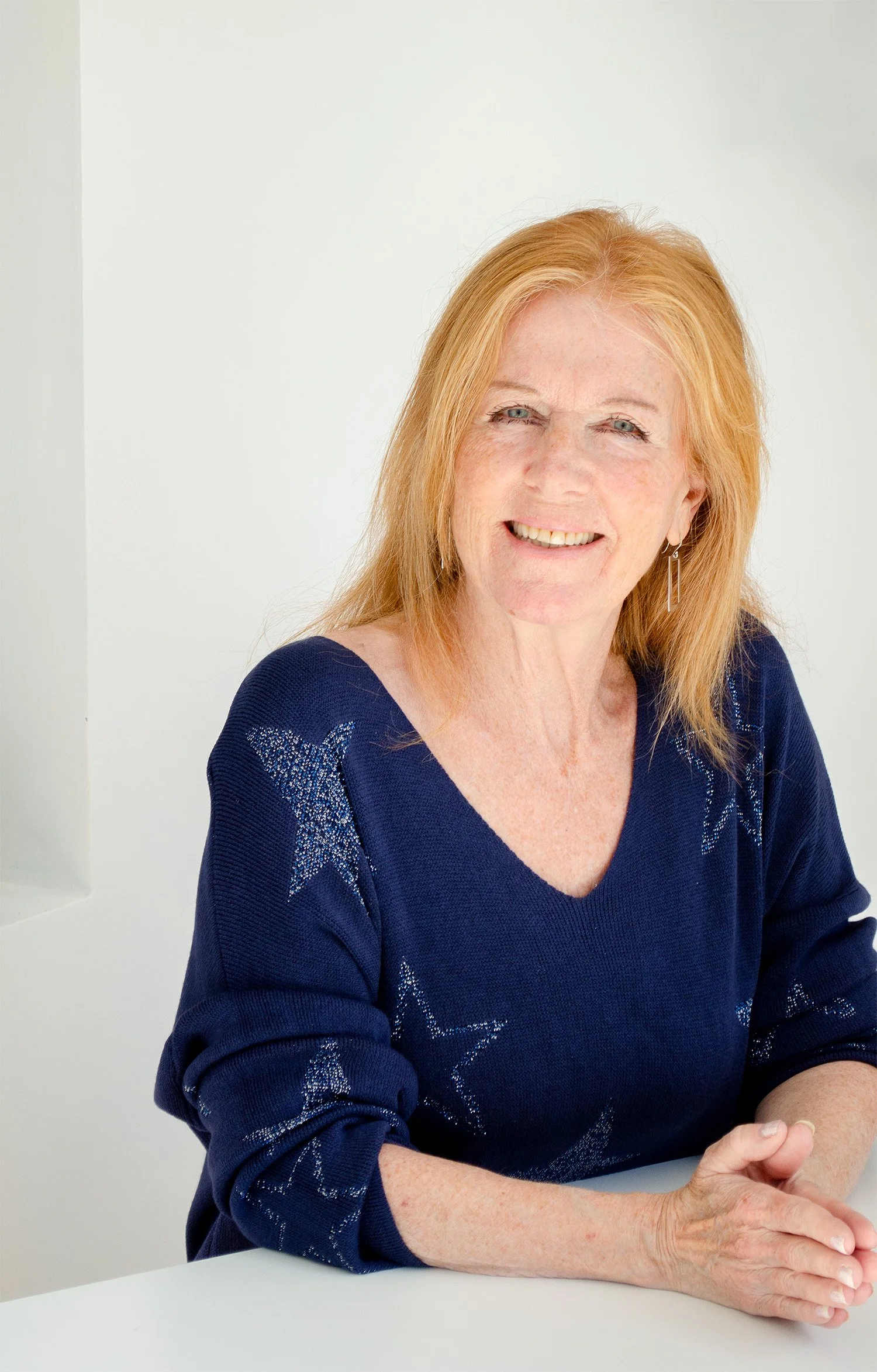 Liz Stokvis wearing a navy blue sweater with star patterns, sitting at a white table with her hands folded.