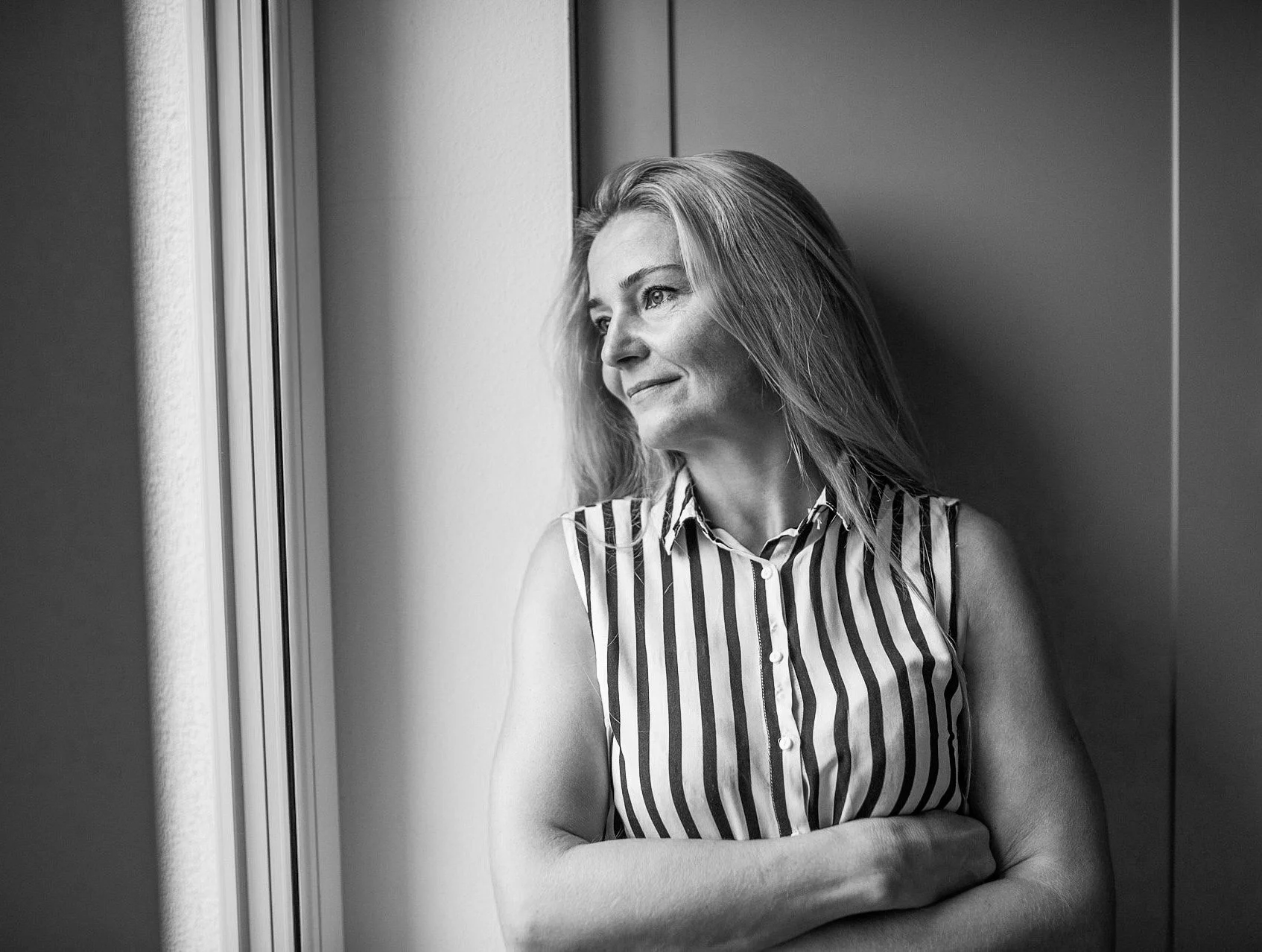 Black and white photo of a woman with light-colored hair, wearing a sleeveless striped shirt, standing by a window, looking out with a thoughtful expression, arms crossed.