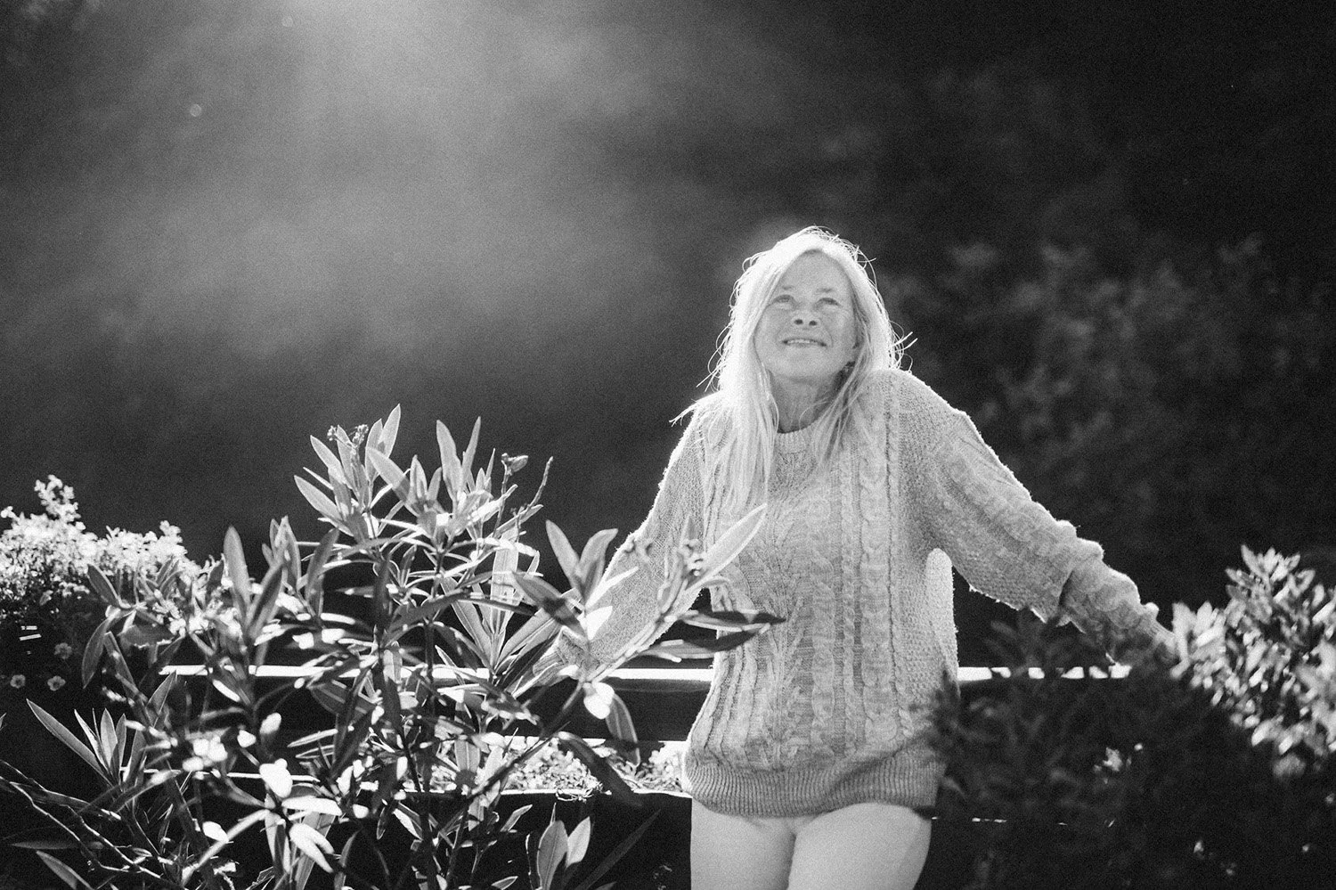 A smiling elderly woman with long light-colored hair, wearing a knit sweater and shorts, standing outdoors among plants at night with a cloudy sky and bright moon in the background.