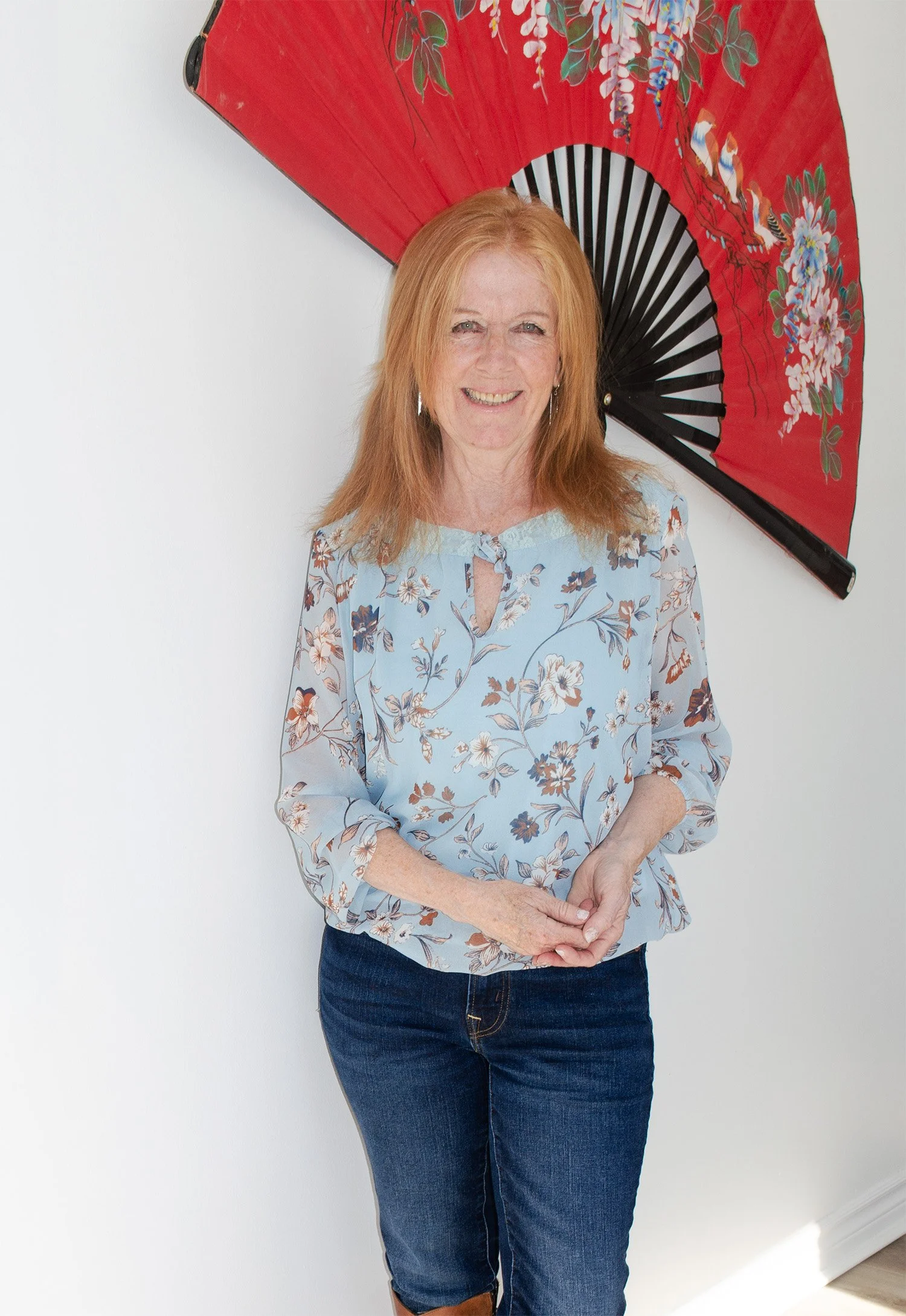 Liz Stokvis coach standing indoors against a plain white wall, smiling, with a large decorative red Asian-style folding fan featuring floral and bird patterns positioned behind her, wearing a light blue floral blouse and dark blue jeans.