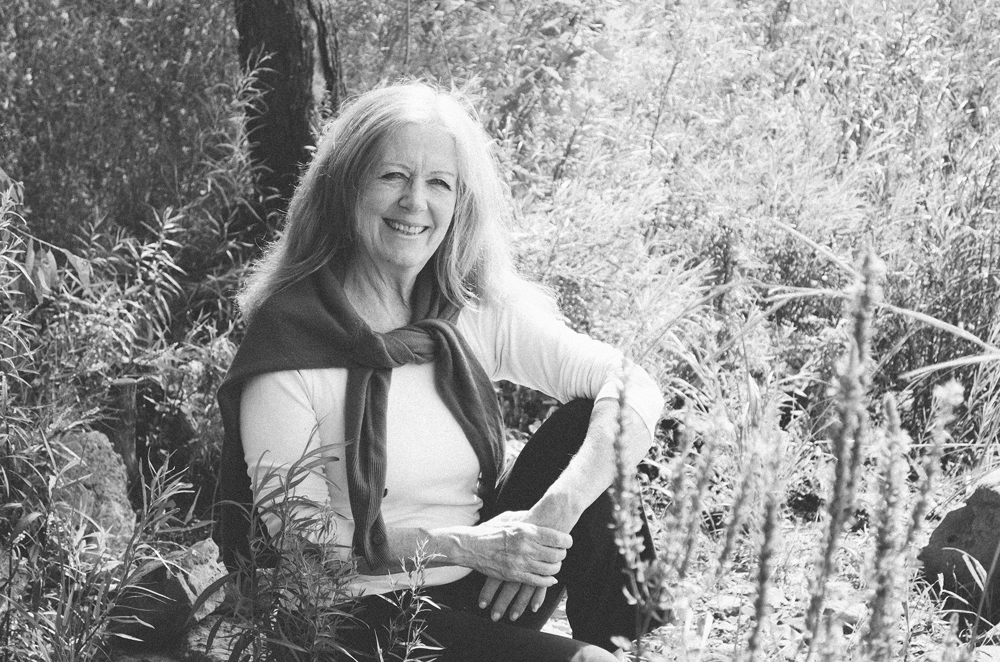Liz Stokvis smiling while sitting outdoors in a natural setting with trees and plants.