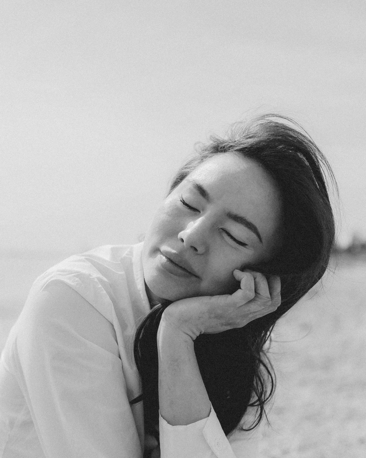 A woman with long dark hair resting her head on her hand with eyes closed, outdoors, black and white photo.