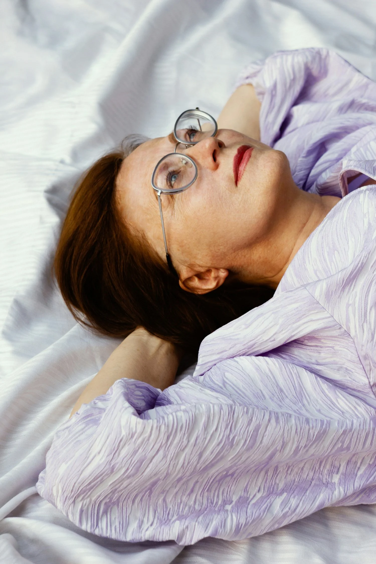 A woman with red hair, glasses, and red lipstick lying on her back on a bed with white sheets, wearing a purple striped pajama top, looking upward.