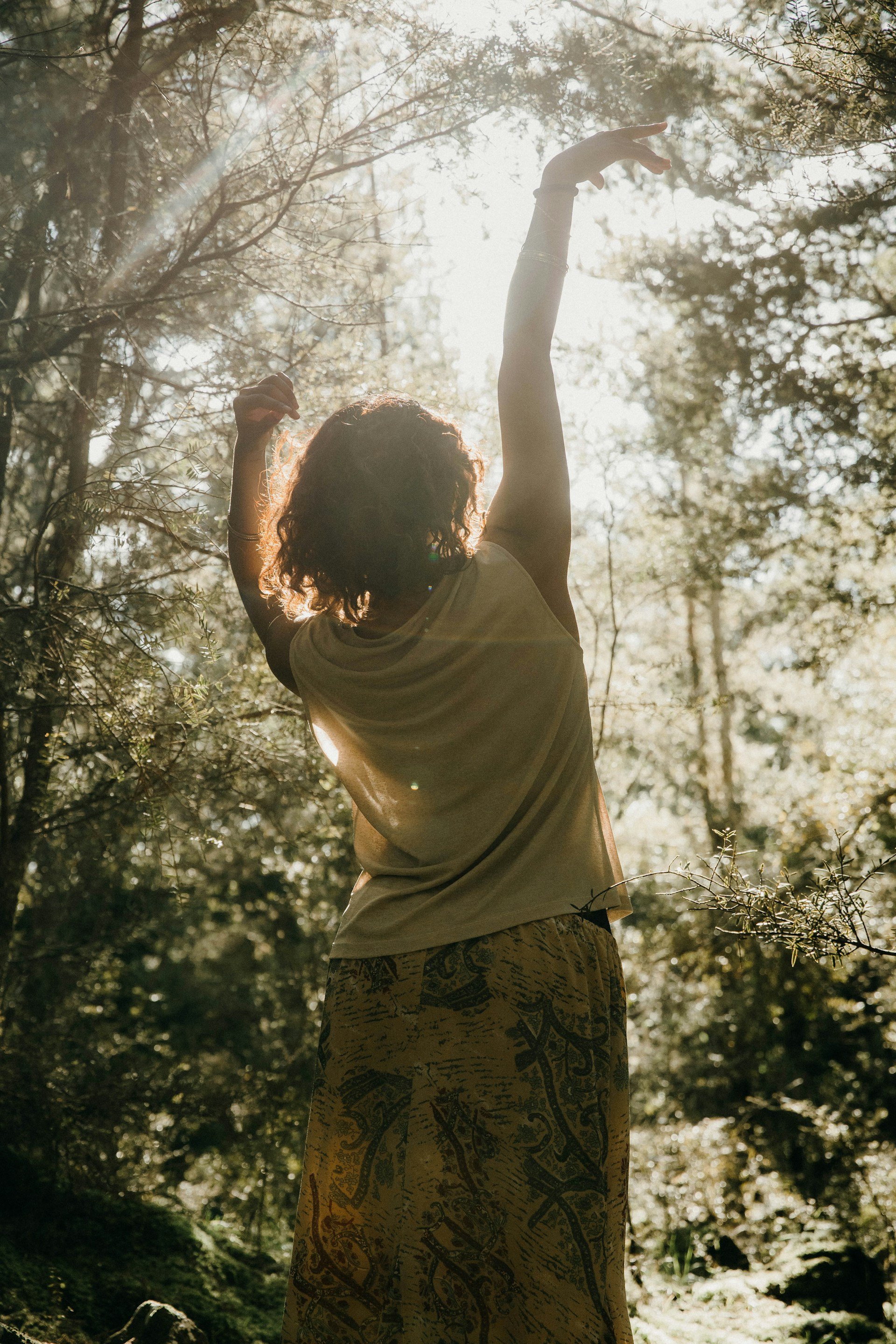 A person with curly hair standing outdoors in a forest, reaching upward with one arm, the sunlight shining through trees.