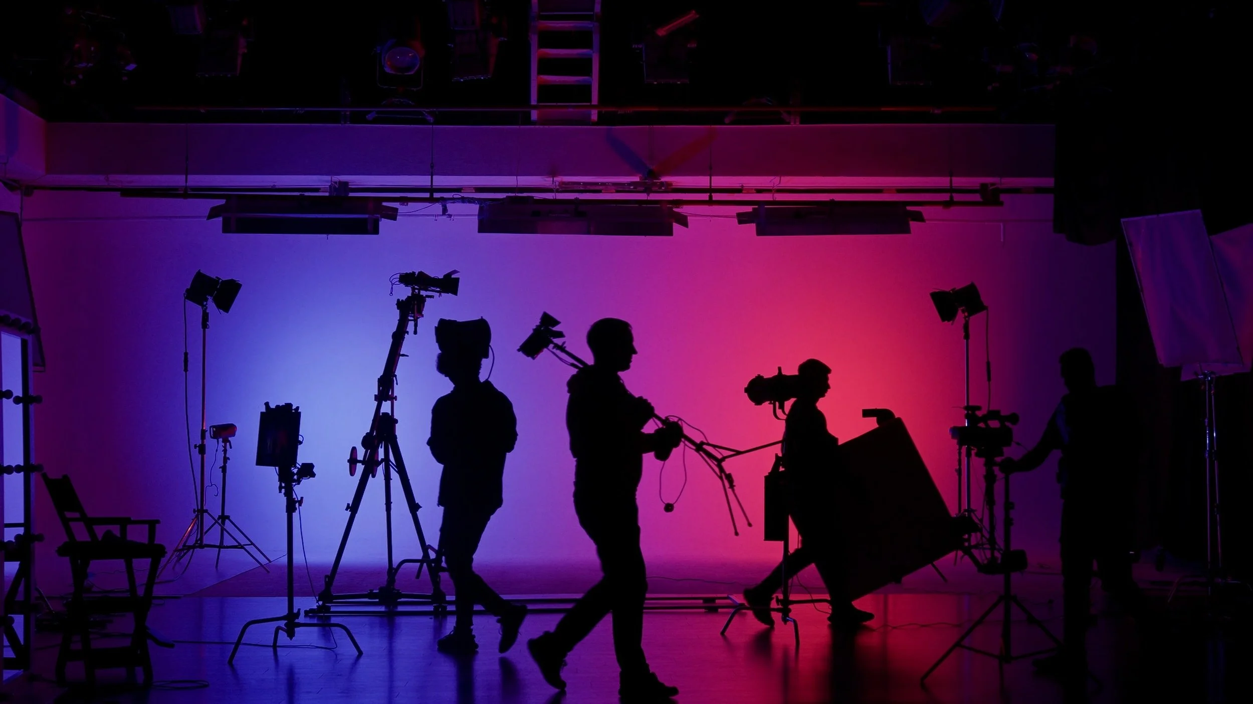 Silhouettes of film crew members working with cameras and lighting equipment in front of a colorful gradient background in a studio setting.