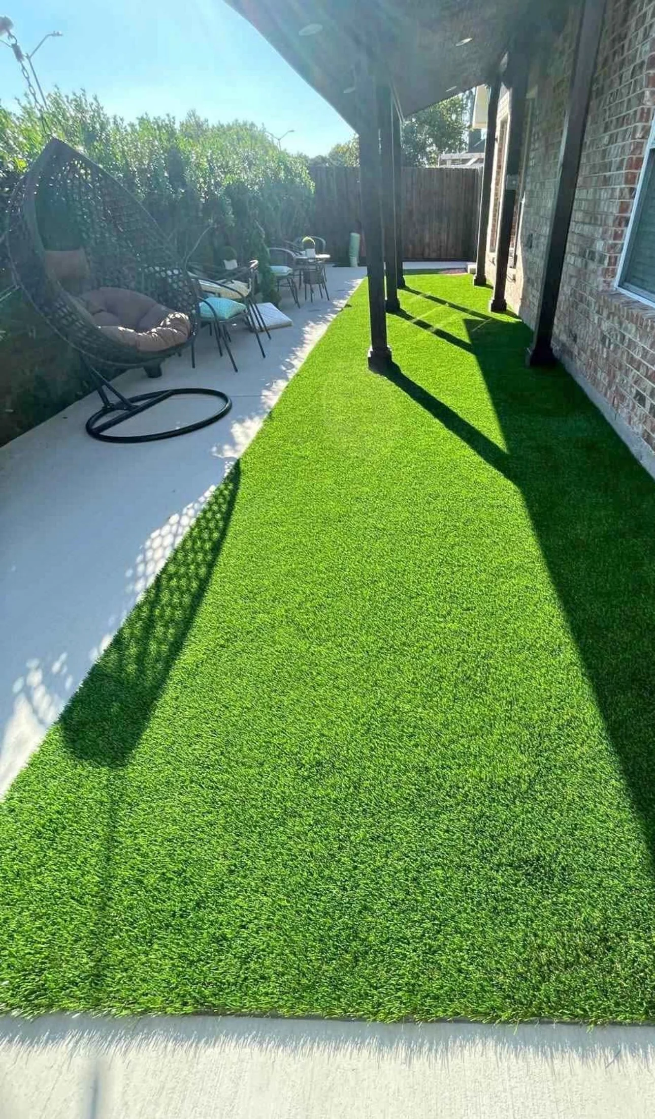 A backyard patio with artificial green grass, outdoor furniture including chairs and a hanging chair, and a brick house with a window.