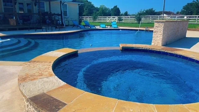 Image of a backyard swimming pool with a hot tub attached, surrounded by a patio with chairs and a white fence in the background.