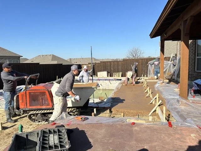 Construction workers pouring concrete on a backyard patio, with a wooden fence and clear blue sky in the background.