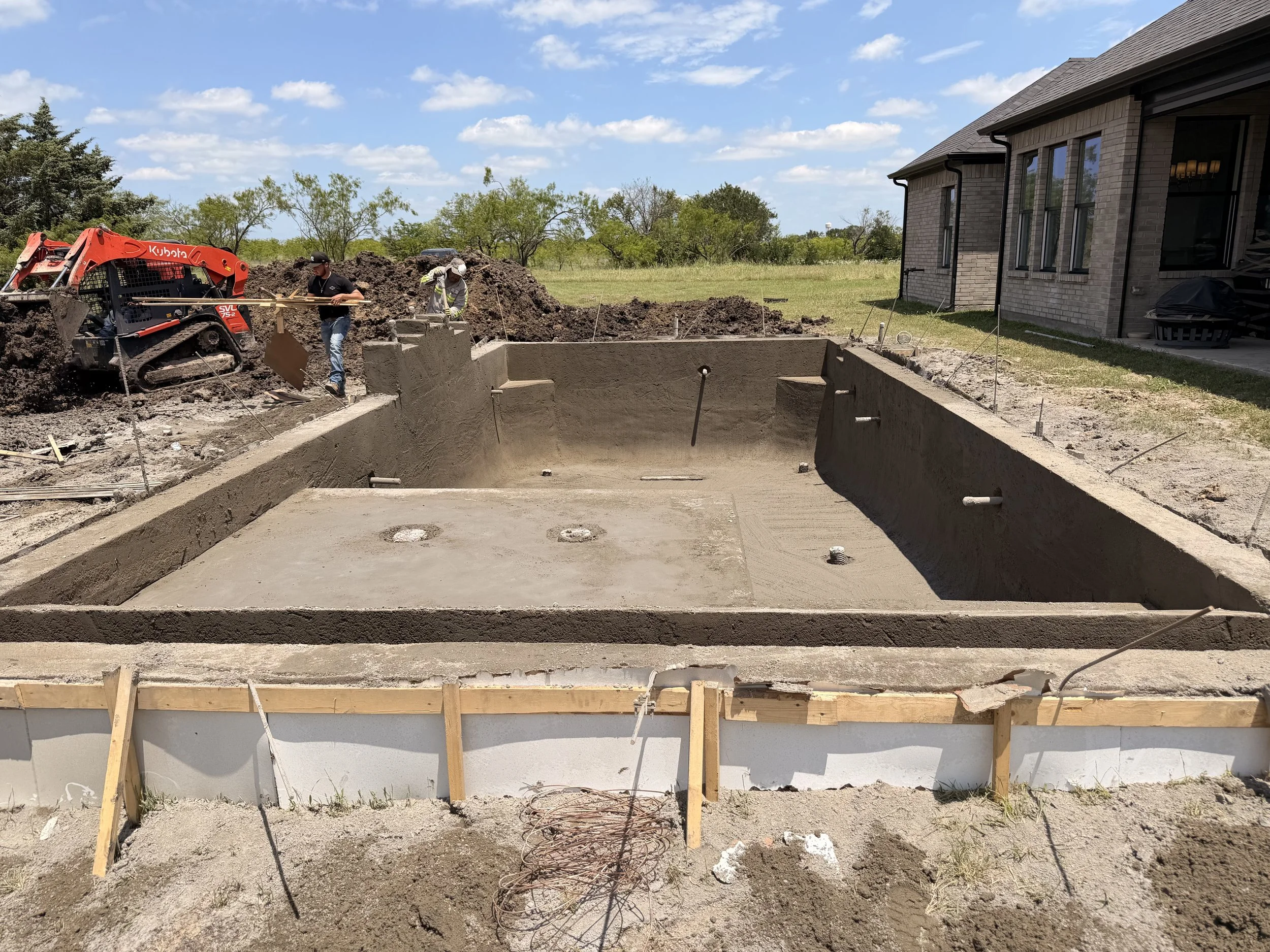Construction site with a concrete foundation, workers, a small excavator, and a partially built house.