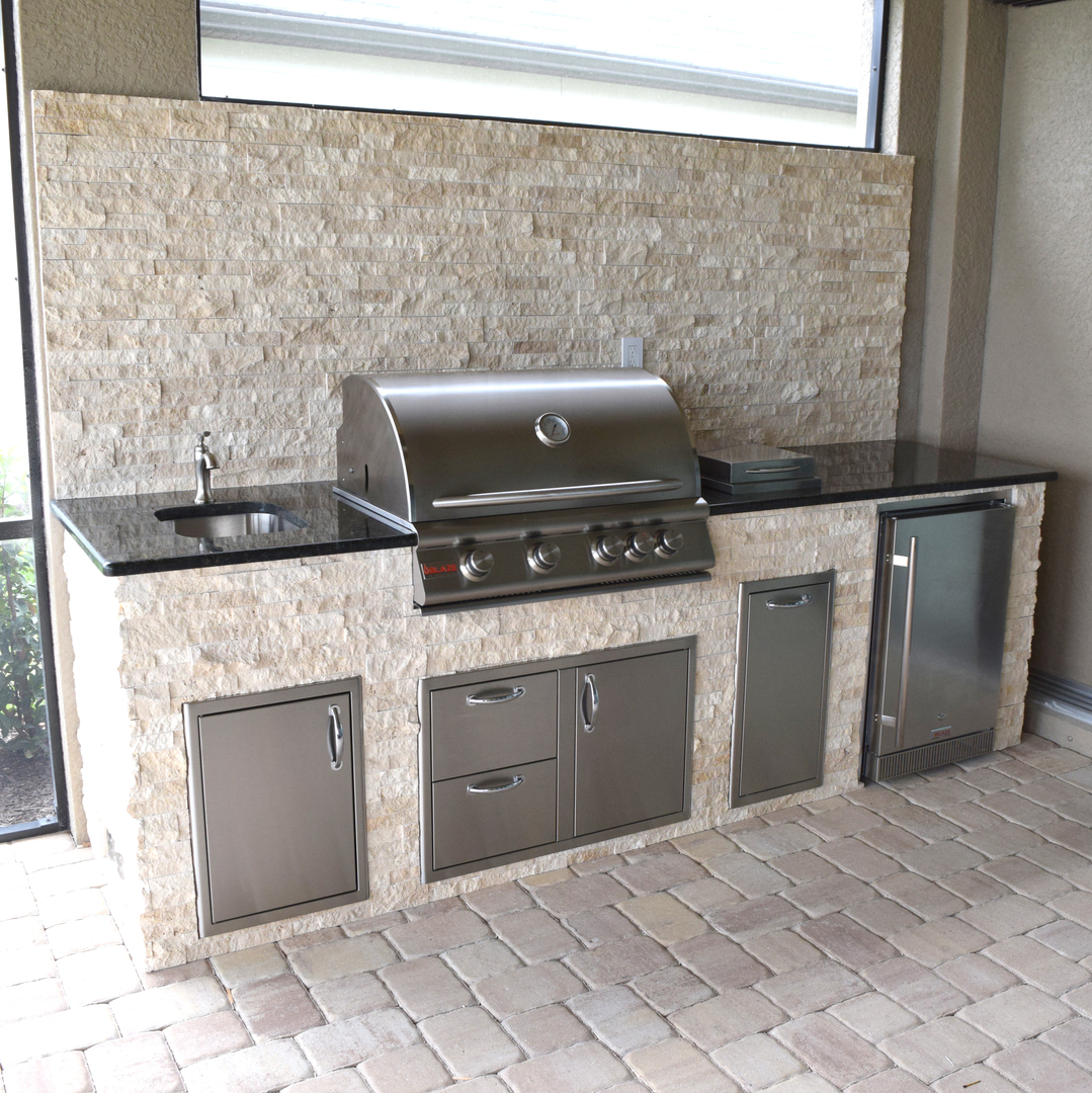 Outdoor kitchen with stainless steel grill, small sink, black countertops, stone wall, and tiled floor.