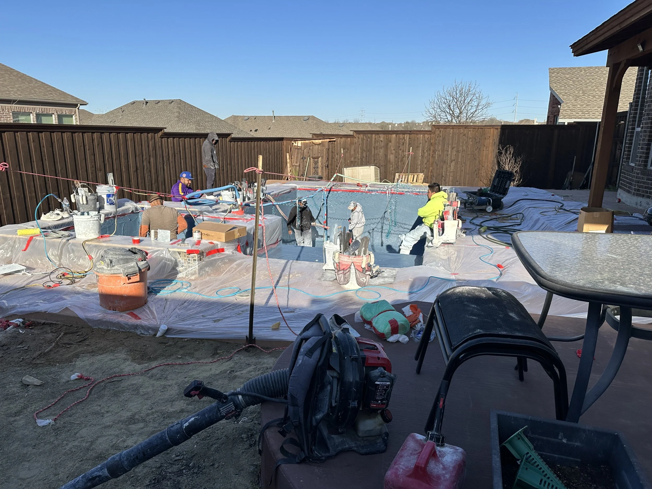 Construction workers installing a swimming pool in a backyard, with some workers standing inside the pool and others around it, surrounded by tools and materials, under a clear blue sky.