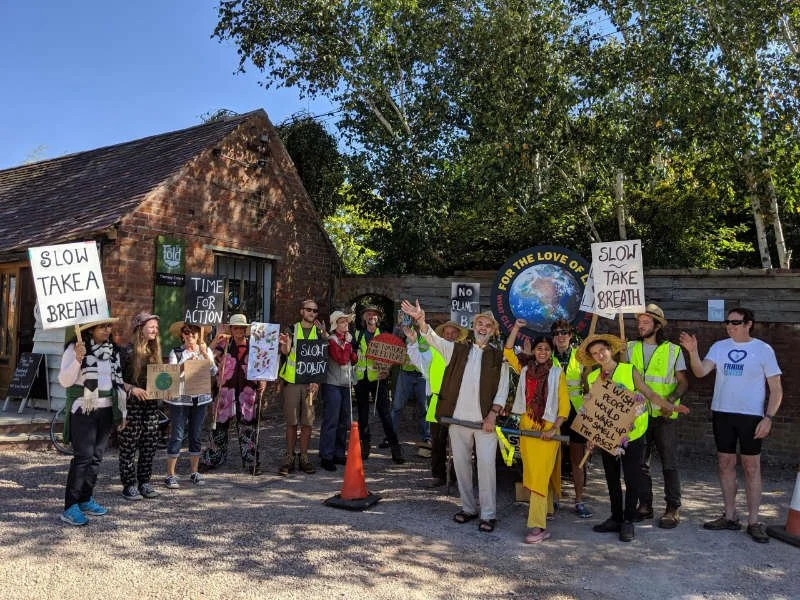 Group of people holding protest signs advocating for climate action and environmental awareness outdoors, with some wearing yellow vests, standing near a small building and trees.