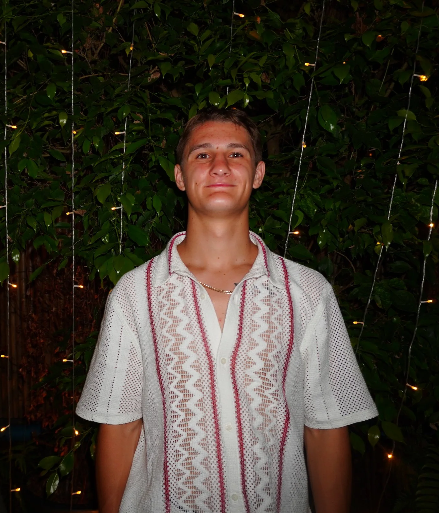 A young man with short hair, wearing a white embroidered shirt, smiling, standing in front of a leafy background with string lights at night.