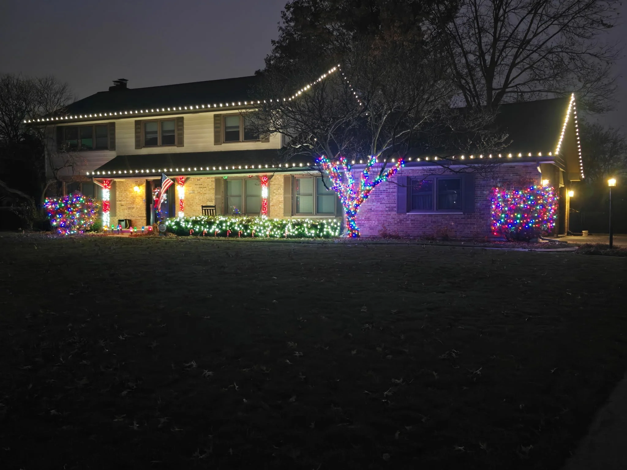 A two-story house decorated with Christmas lights, including strings of white lights along the roofline, a tree in front wrapped with multicolored lights, and bushes decorated with multicolored string lights. The house has three visible windows and a front porch with some holiday decorations. The scene is set at night.