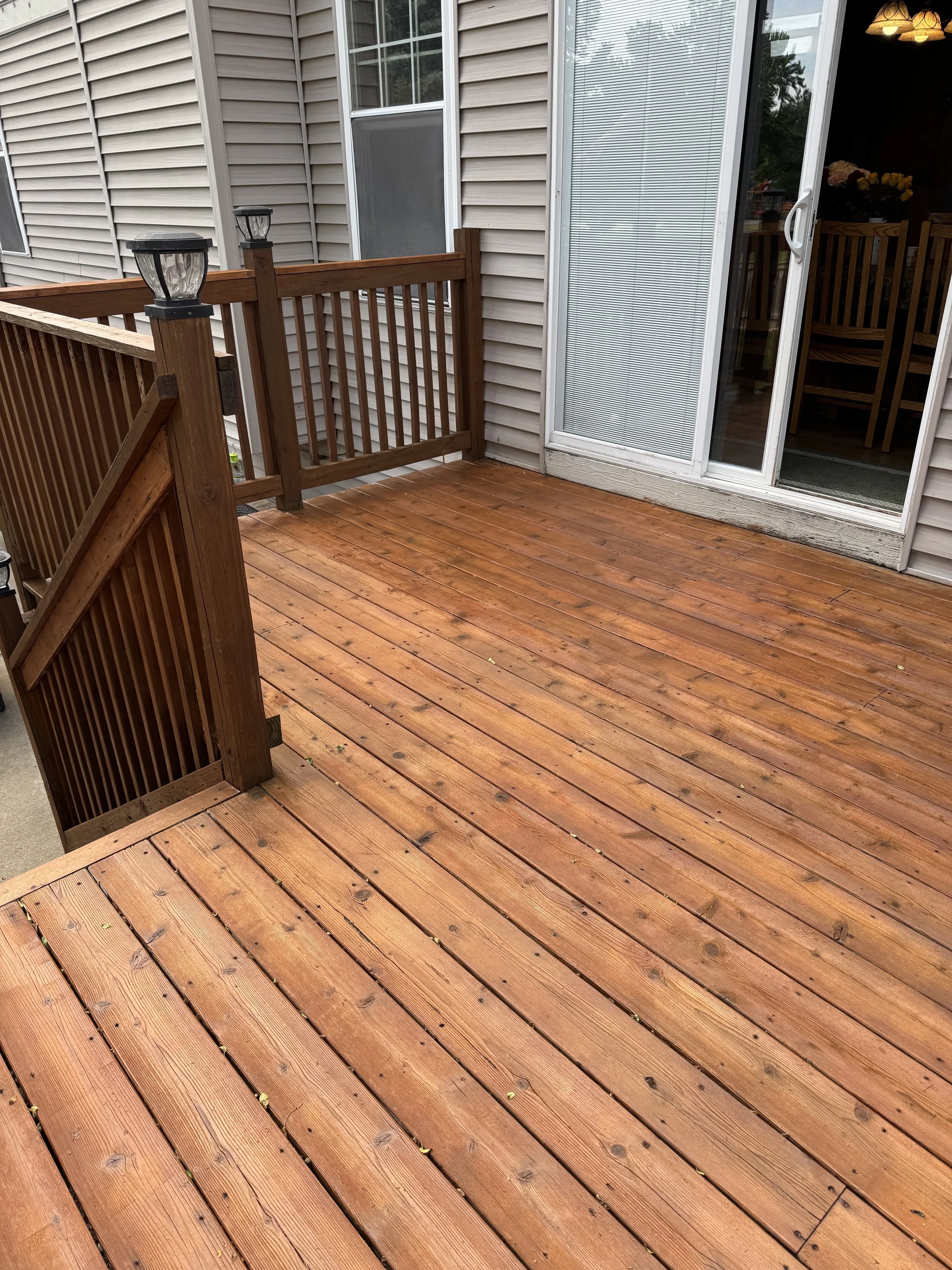 A wooden deck attached to a house with sliding glass doors and an interior visible through the door. The deck has wooden railings with light fixtures on top. The house exterior features beige vinyl siding and a window with blinds.