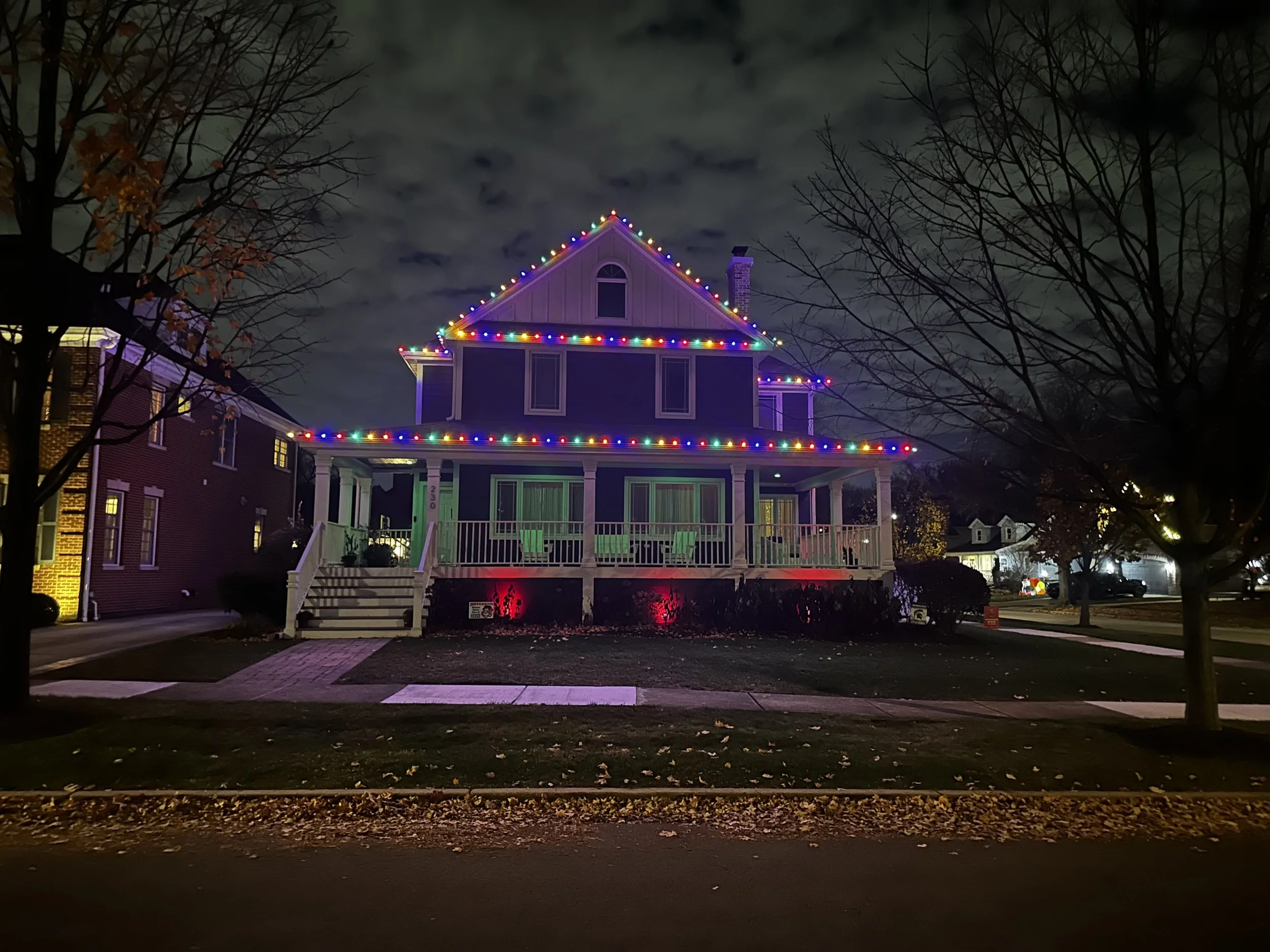 A two-story house decorated with colorful Christmas lights at night, with leafless trees in the foreground and a cloudy sky in the background.