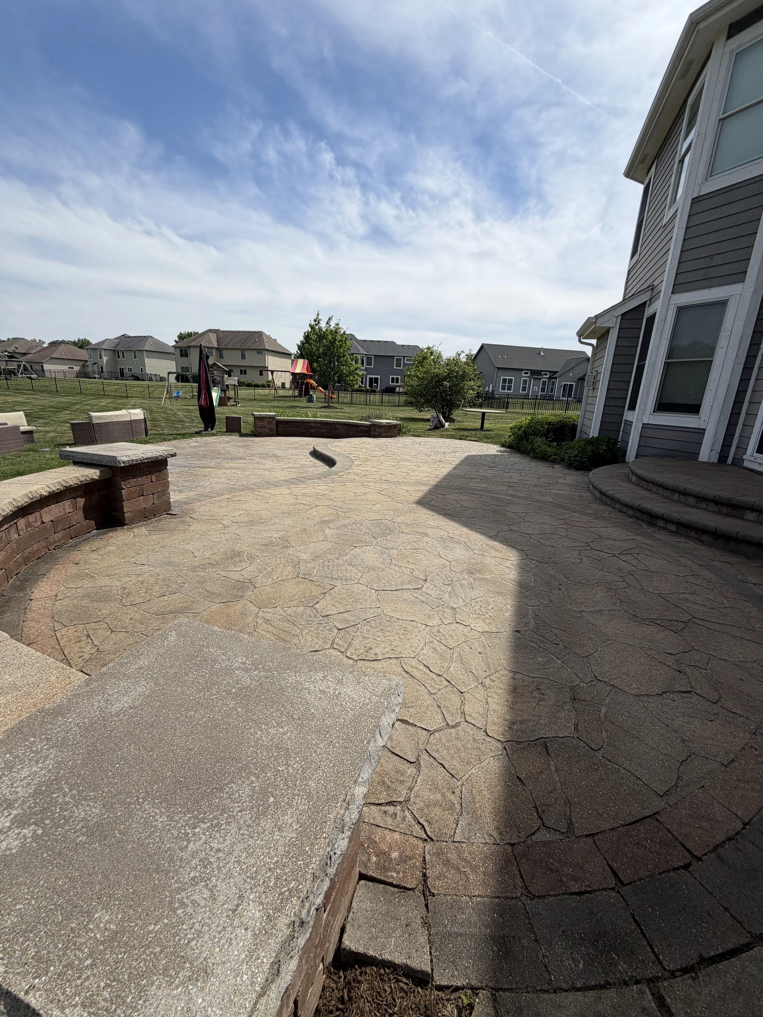 View of a backyard patio with a stone and concrete surface, a curved brick wall, grass lawn, trees, and neighboring houses under a partly cloudy sky.