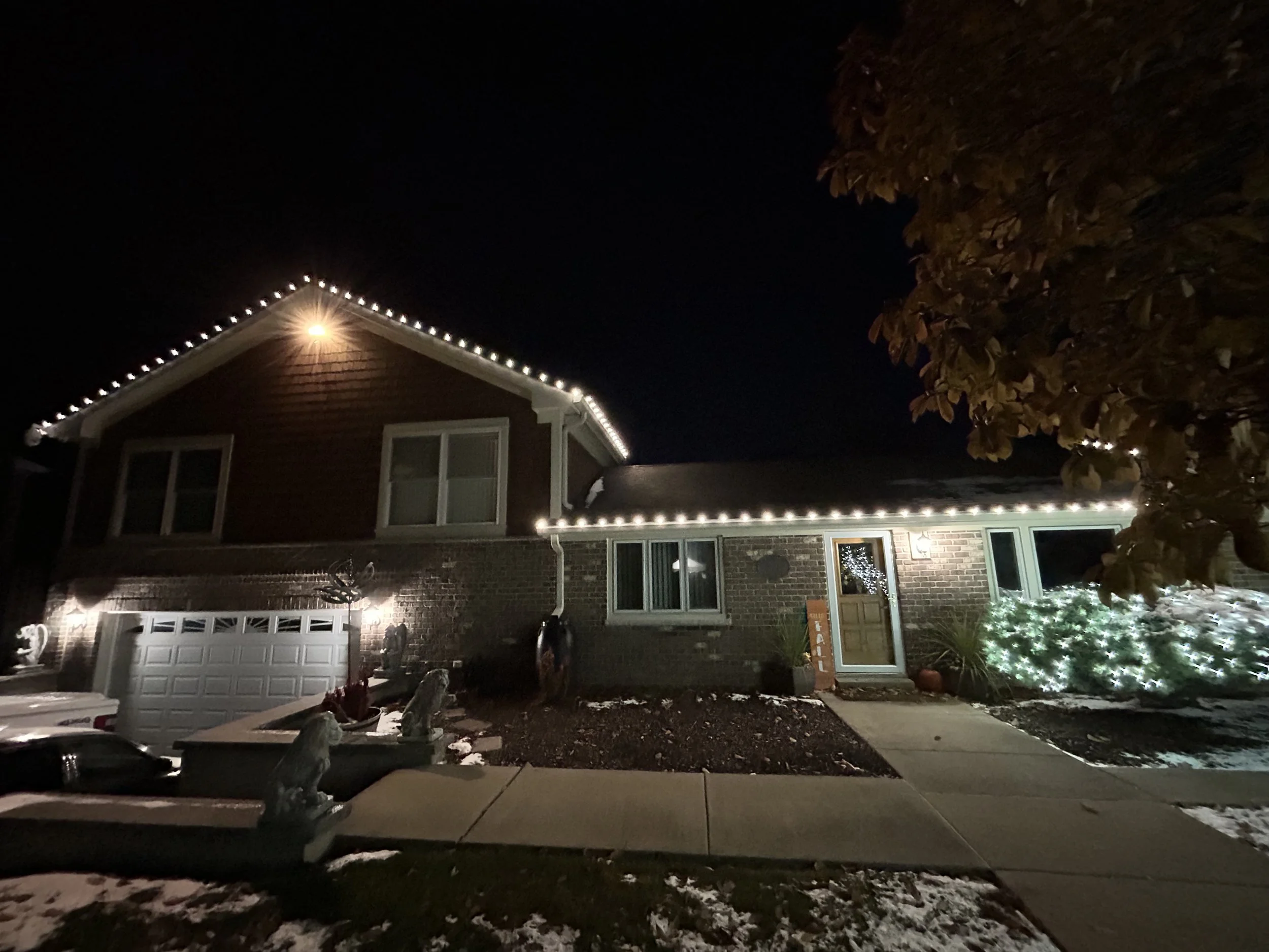 A house decorated with white Christmas lights outline the roof, and a small lit Christmas tree is visible near the front door. There is some snow on the ground and on a bench in the yard. A tree with brown leaves is on the right side of the image, partially blocking the view of the house. The house has a brick and siding exterior with multiple windows.