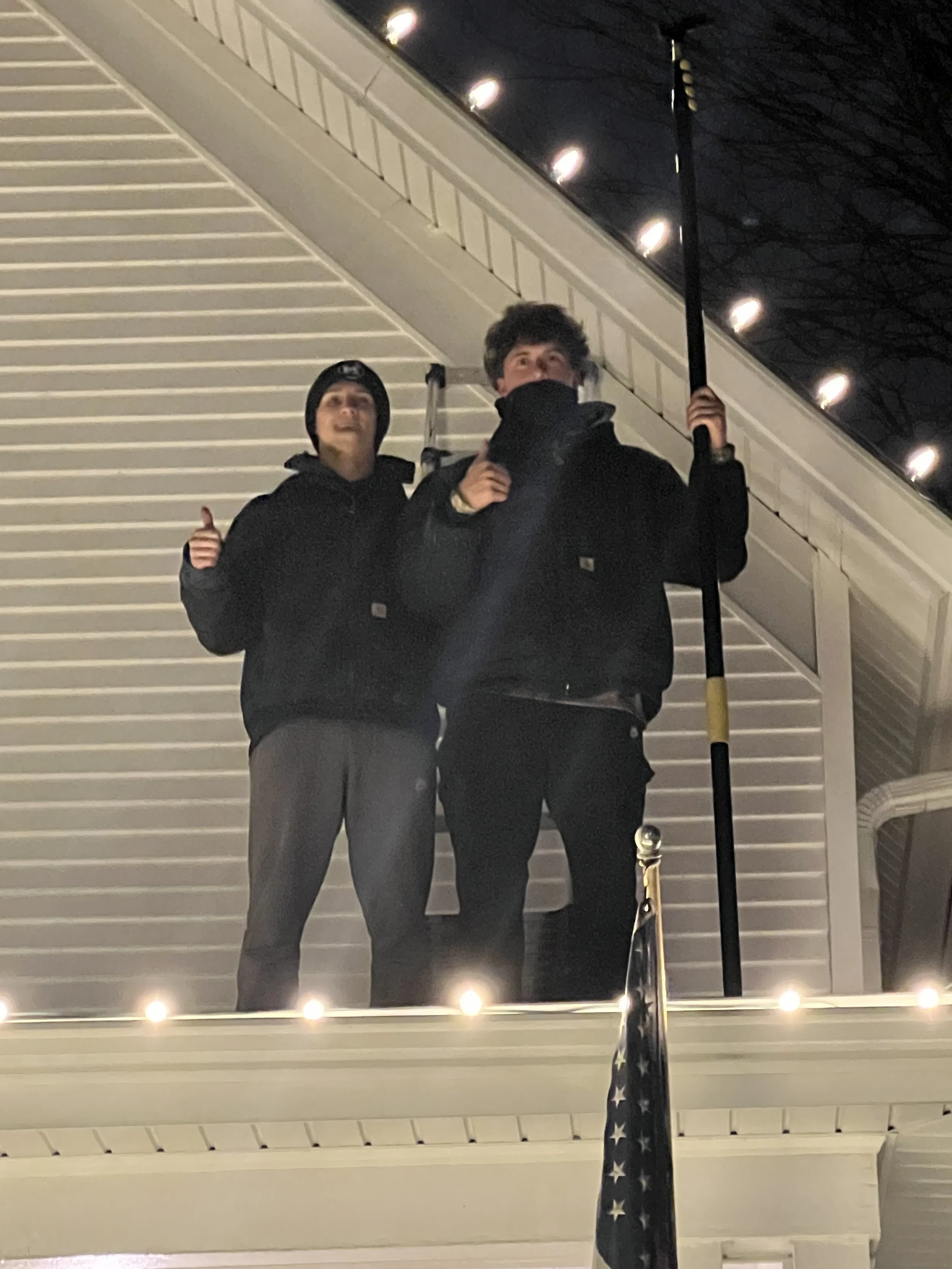 Two young men standing indoors in front of a mirror, one giving a thumbs-up and the other holding a flagpole with an American flag. Both are wearing black jackets and dark pants, and the background features white siding and ceiling lights.