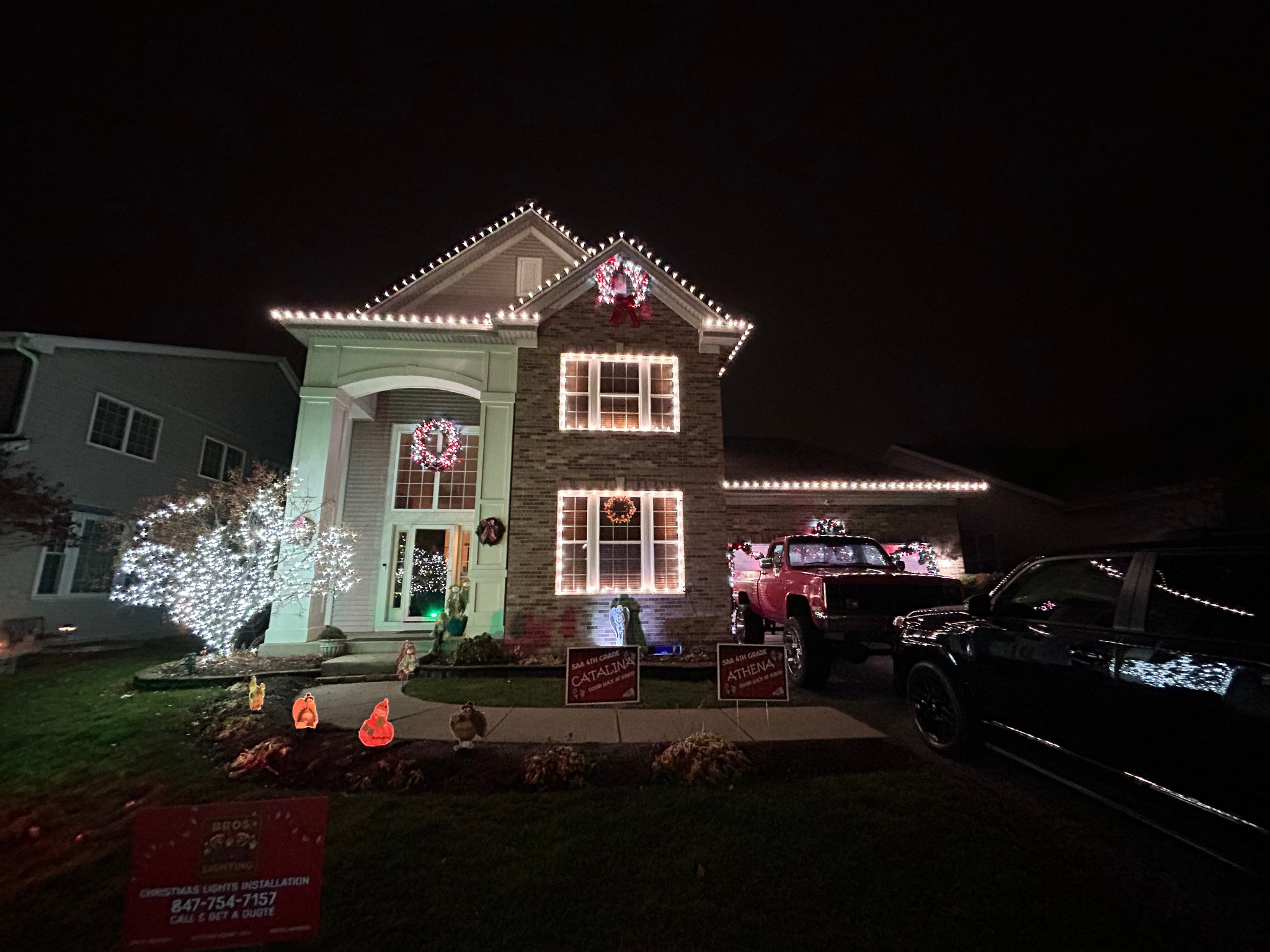 A two-story house decorated with Christmas lights, wreaths, and Christmas figures in the yard at night. Multiple cars are parked in the driveway.