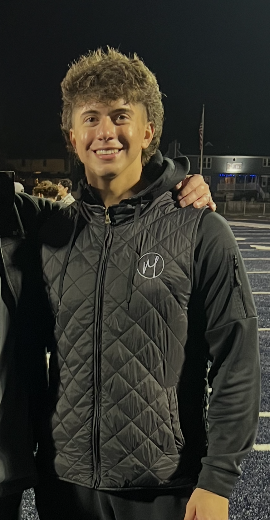 A young man with curly brown hair smiling at night, wearing a black quilted jacket with a circular logo on the chest, standing in a parking lot with some people and buildings in the background.