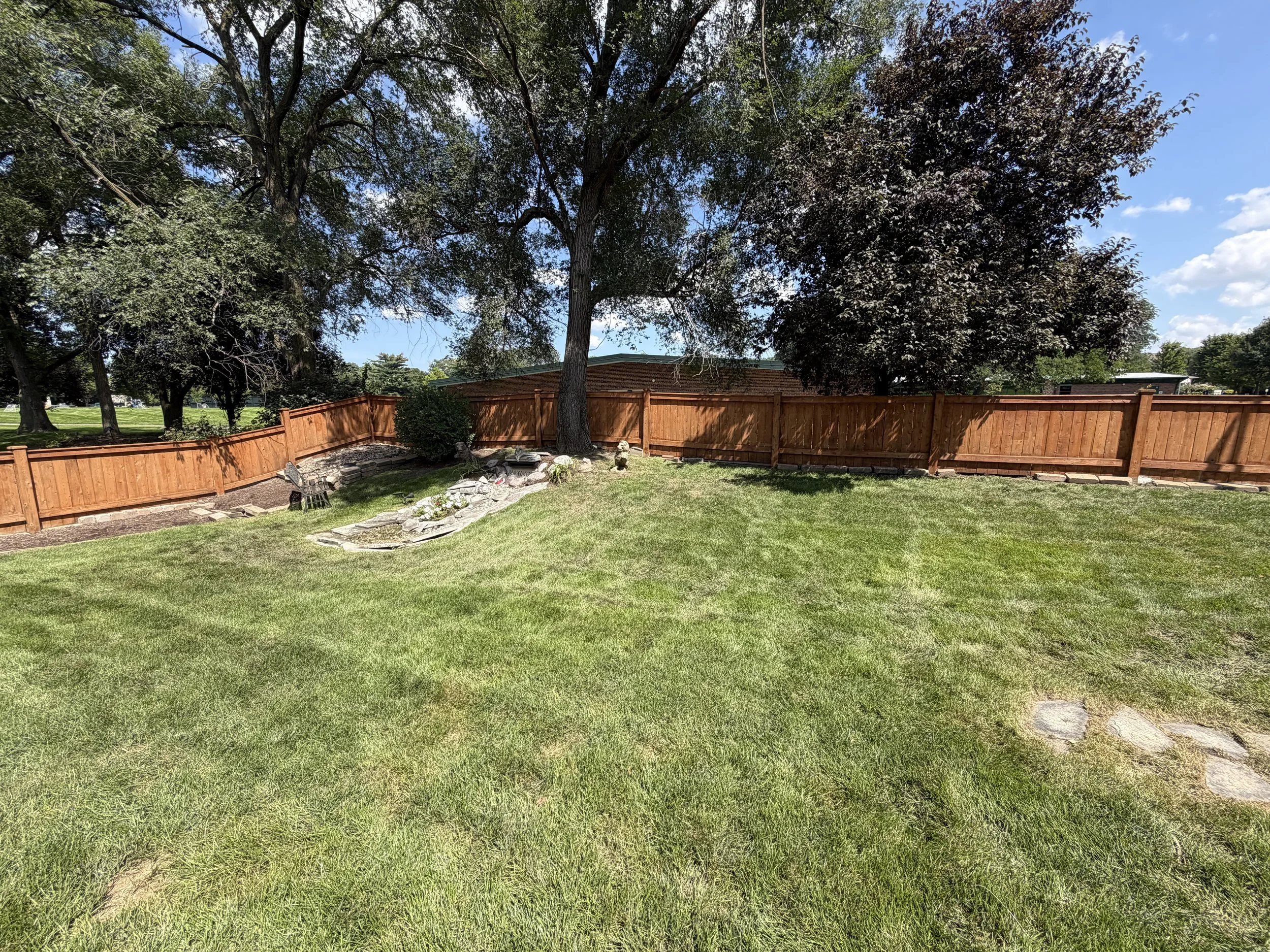 A backyard with a green lawn, a wooden fence, several large trees, and a small stone pathway. The sky is mostly clear with a few clouds.