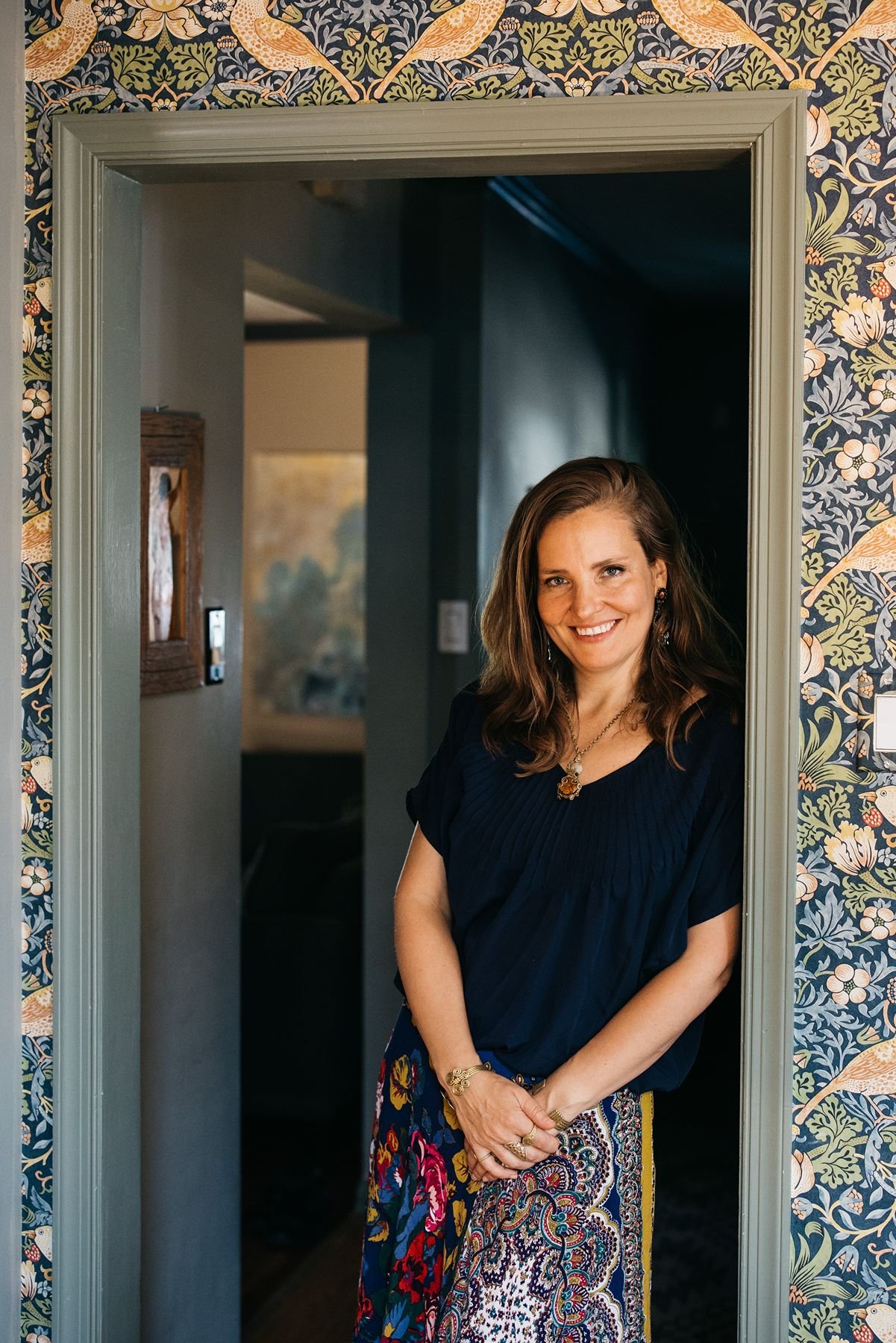 A woman standing in a doorway framed by floral wallpaper, smiling at the camera. She is wearing a dark top and colorful, patterned skirt, with jewelry including earrings, a necklace, and rings.