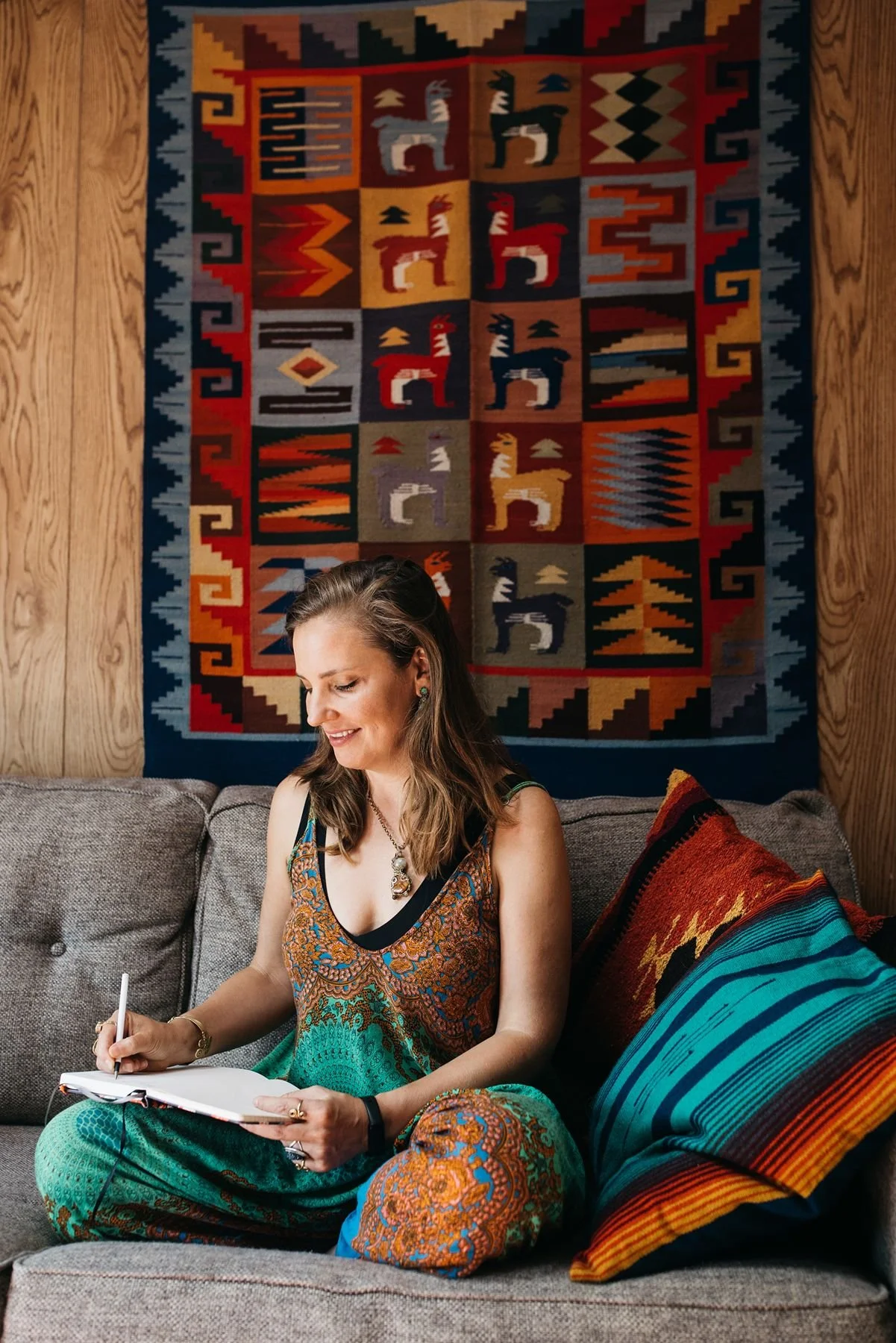 A woman sitting on a couch, writing in a notebook, with colorful patterned pillows and a woven tribal-style wall hanging in the background.