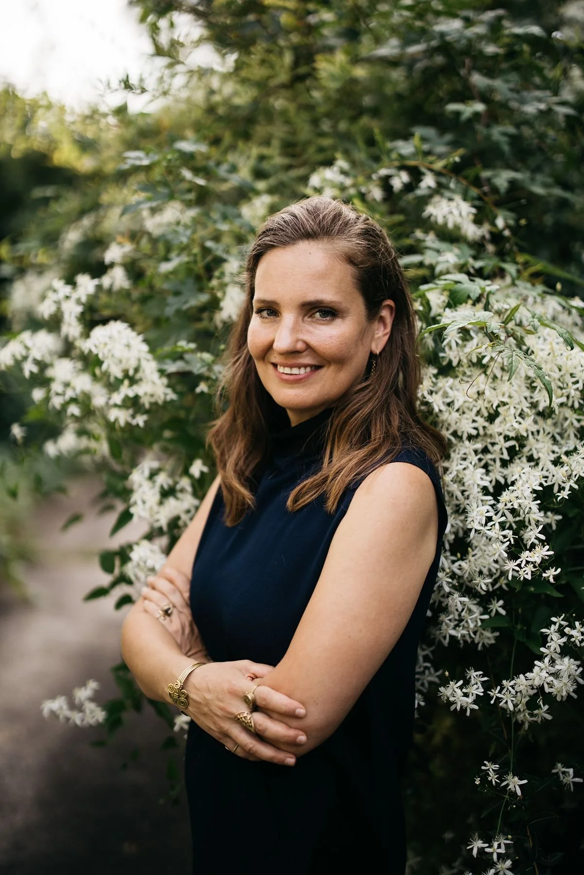 A woman with long brown hair wearing a sleeveless black dress and gold jewelry standing outdoors in front of a bush with white flowers, smiling, with arms crossed.