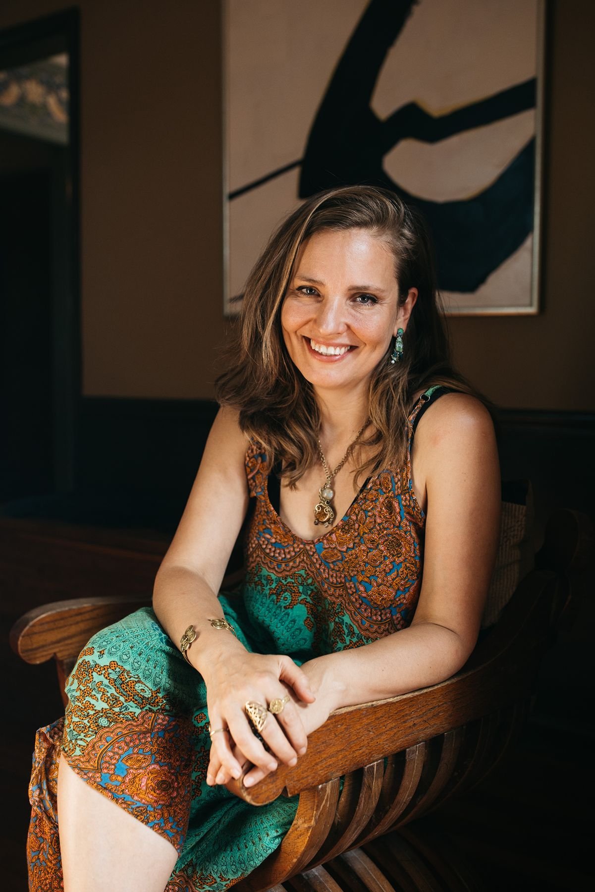 A woman with light brown hair smiling, wearing a patterned dress and jewelry, sitting on a wooden bench in a dimly lit room with a large abstract black and beige painting in the background.