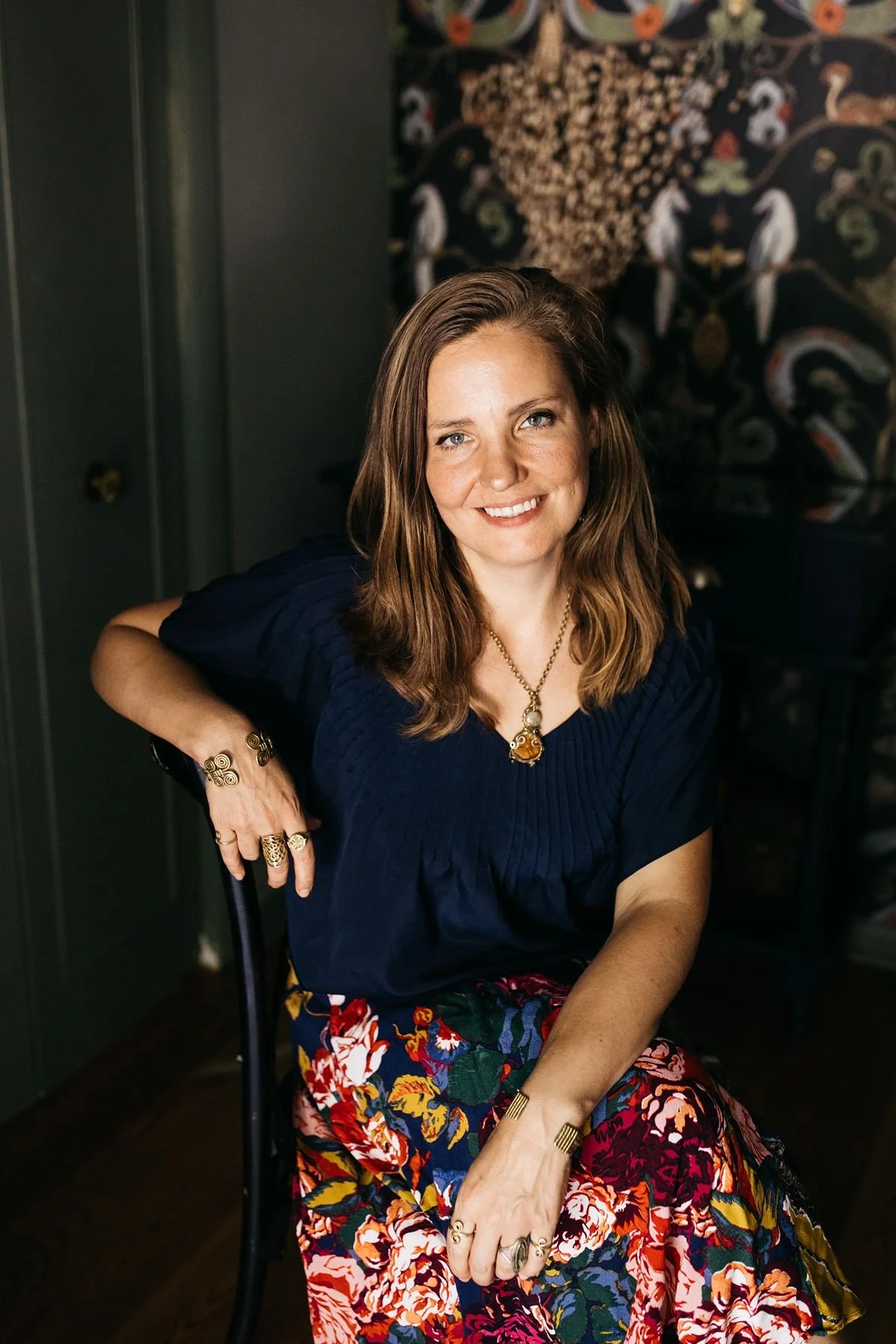 A woman with light skin and long brown hair, wearing a dark blue top, floral skirt, and gold jewelry, sitting indoors with a patterned tapestry in the background.