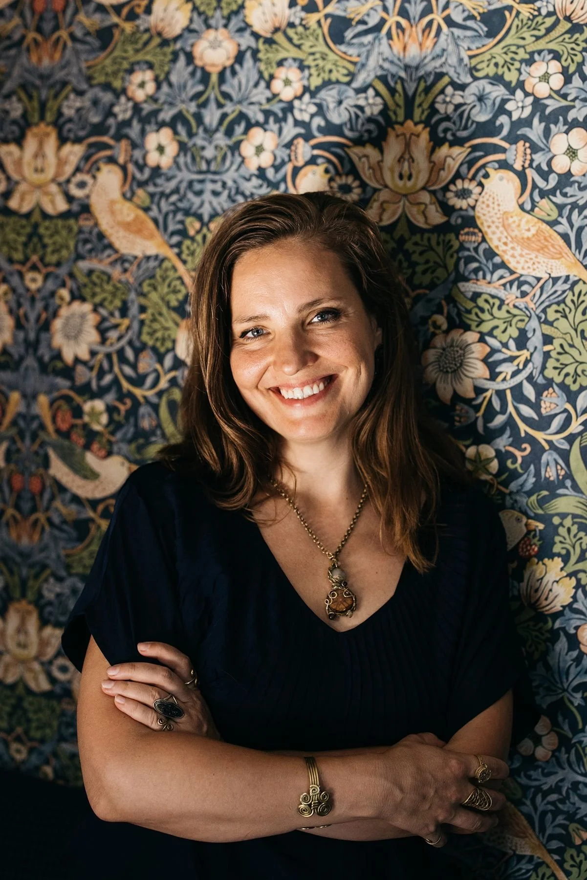 A woman with shoulder-length brown hair smiling, wearing a dark top and jewelry, standing in front of a patterned wallpaper with birds, flowers, and leaves.