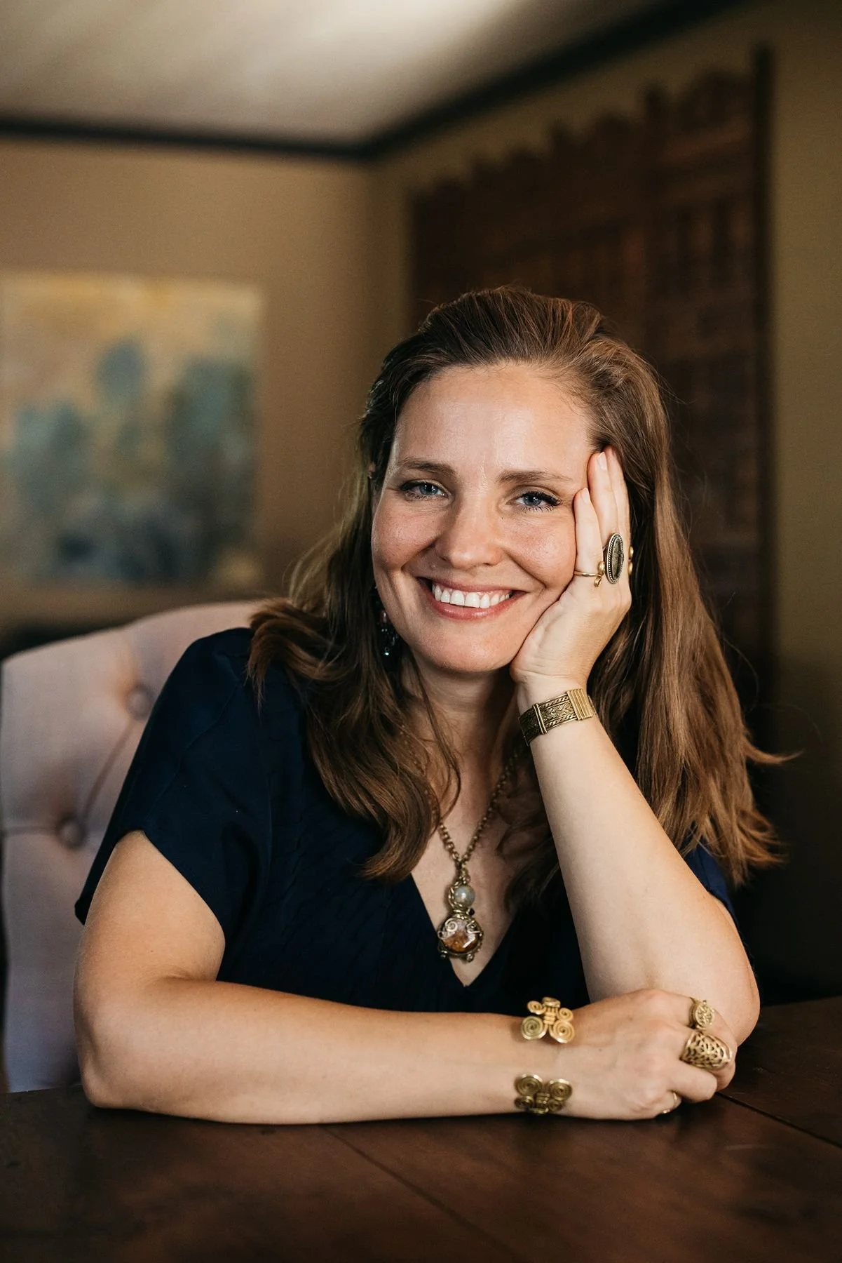 A woman with long, wavy brown hair, smiling, sitting at a table with her head resting on her hand. She is wearing a dark blue top and gold jewelry, including rings, bracelets, a necklace, and earrings. The background features a blurred painting and a decorative wooden piece.