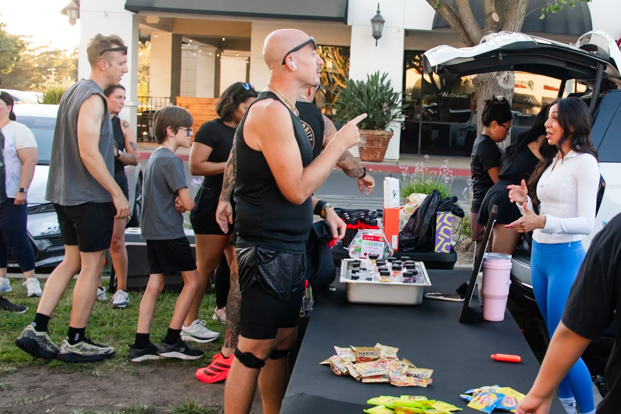 A group of people outdoors in line at a vendor table, engaging in conversation; the vendor, a woman in a white shirt, is talking to a customer, a man with a shaved head and tattoos on his arms. Items on the table include snacks and small products. Ot