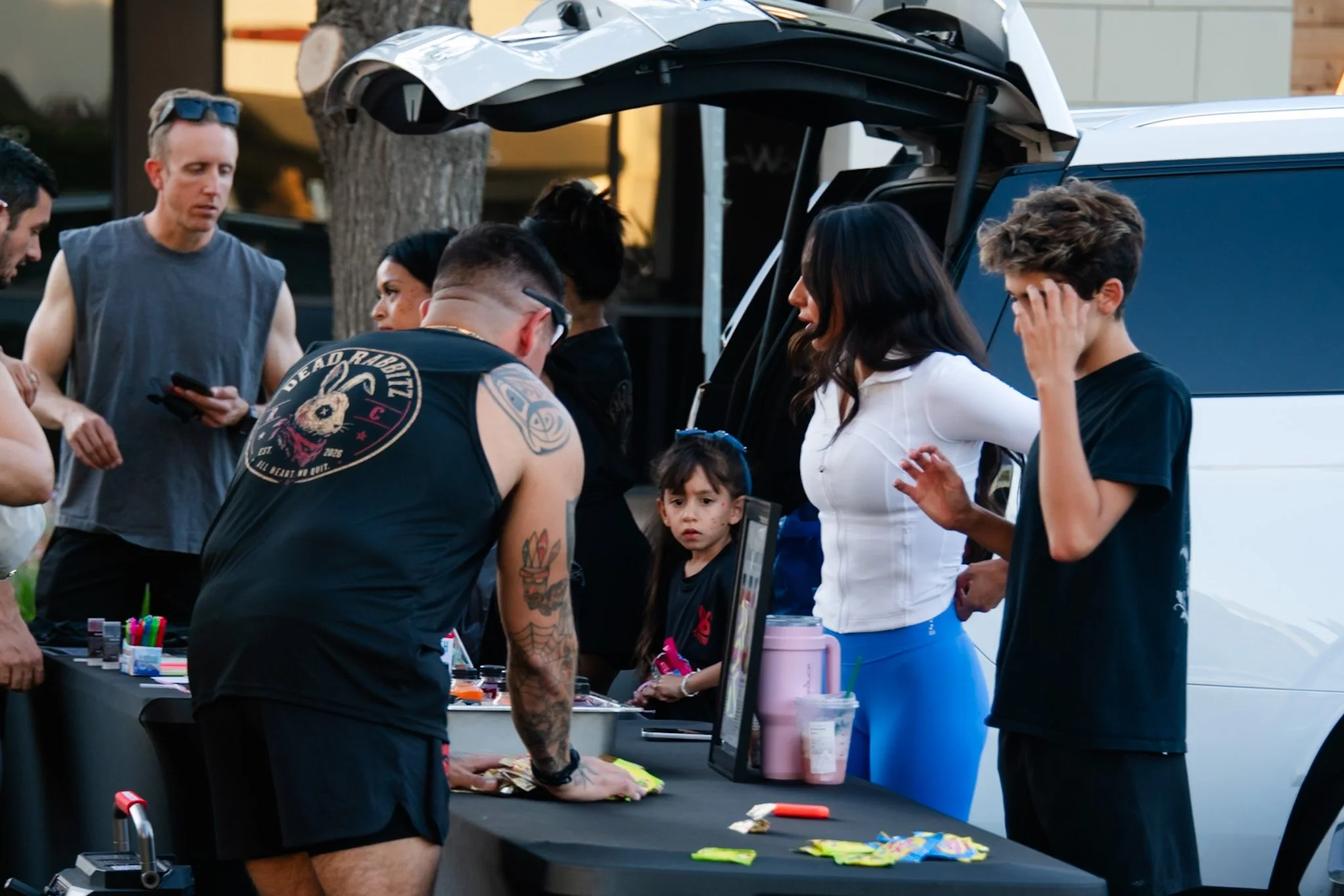 A group of people gathered around a table outdoors, engaging in a discussion or activity. There are various colorful items on the table, and some individuals are looking at handheld devices or examining objects. The setting appears to be casual, with
